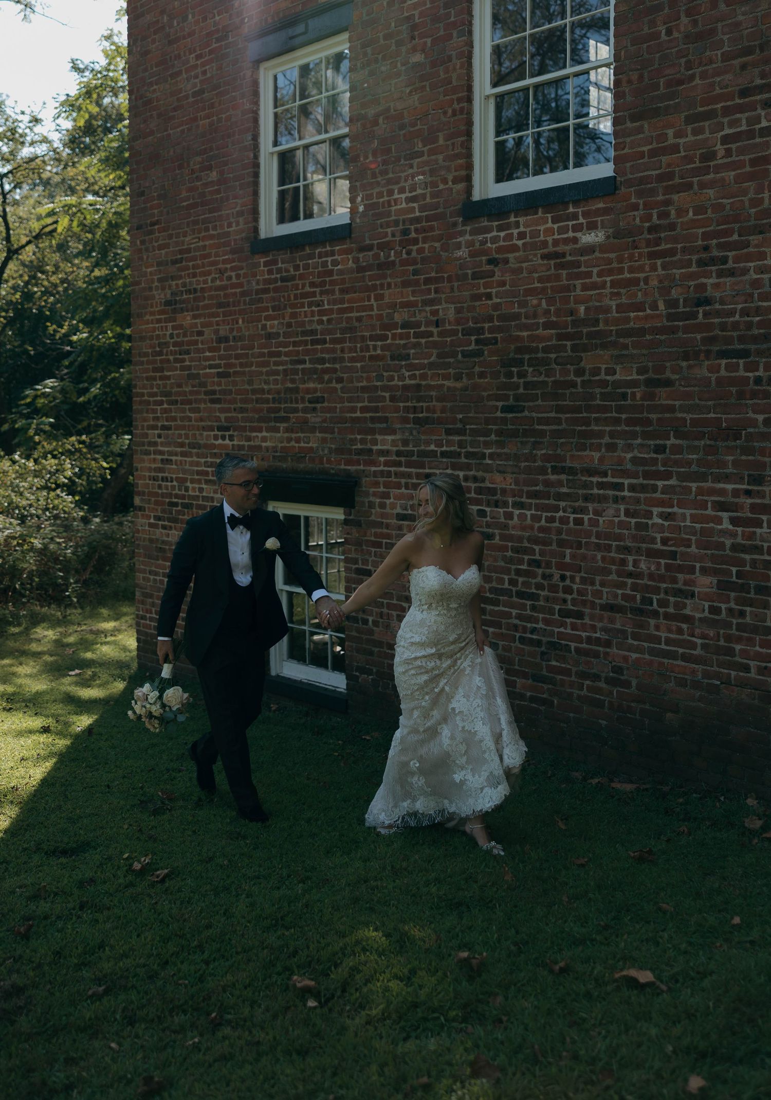 A wedding couple walks beside a historic red brick building on a sunny afternoon.