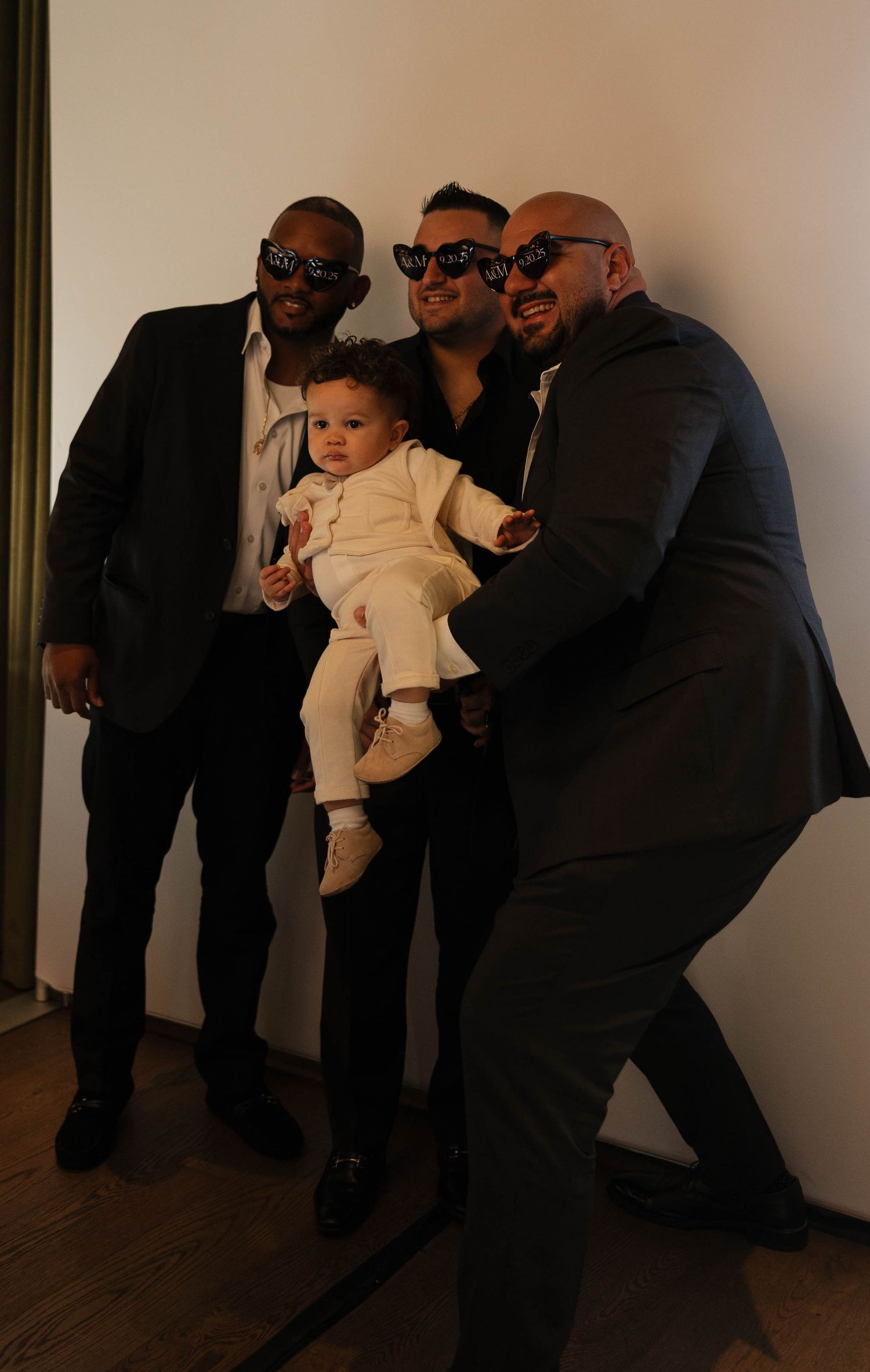 Groomsmen in suits and sunglasses pose together while holding a baby in formal wear.
