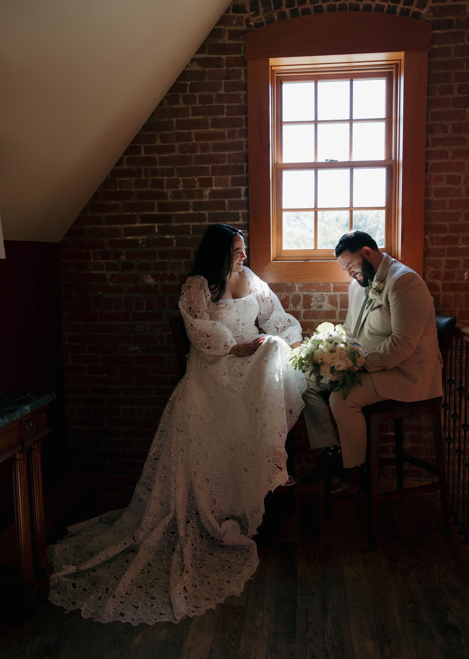 Wedding couple poses in a rustic brick attic space with dramatic window lighting.