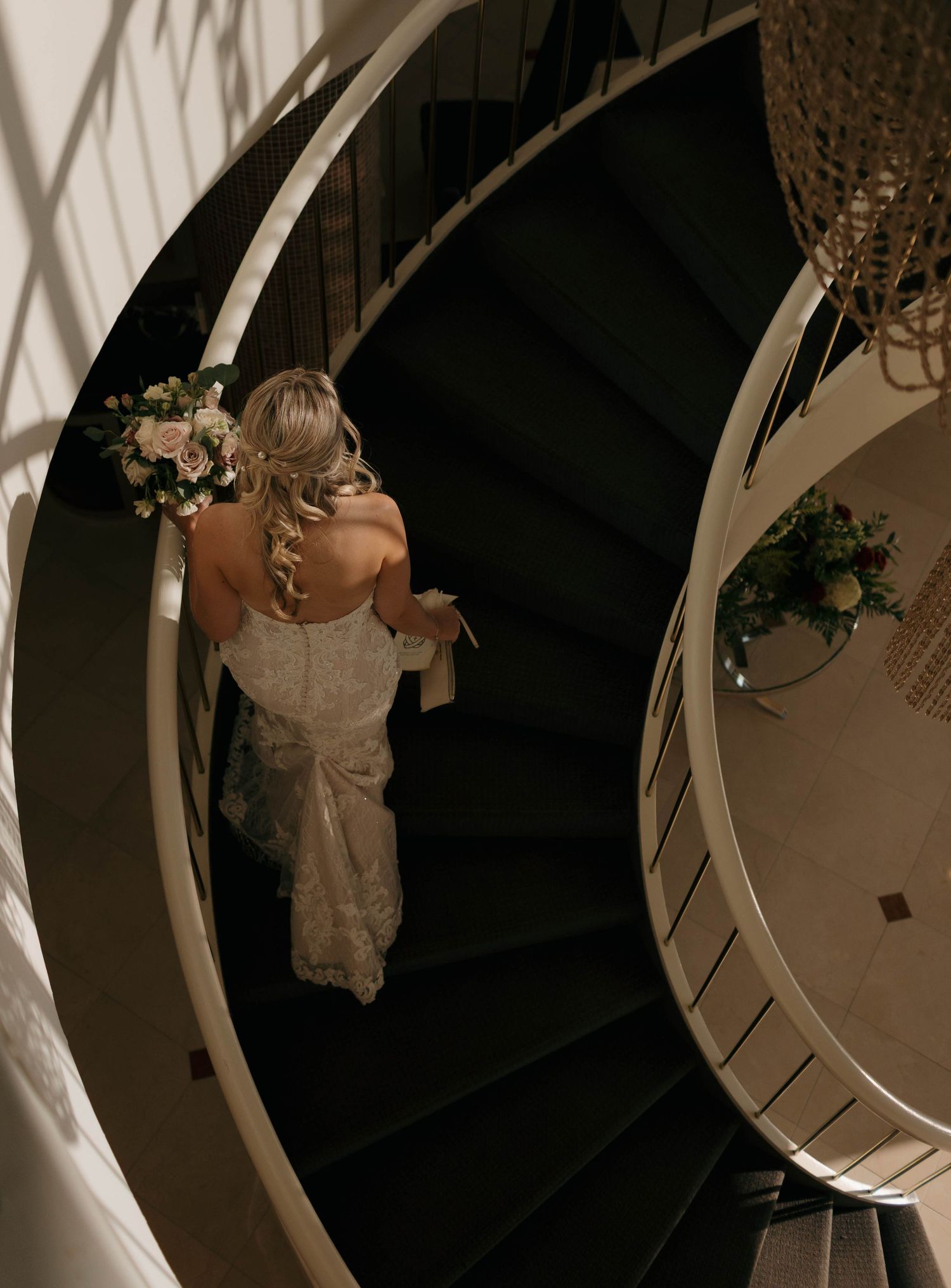 A bride in a lace gown descends an elegant spiral staircase holding a floral bouquet.