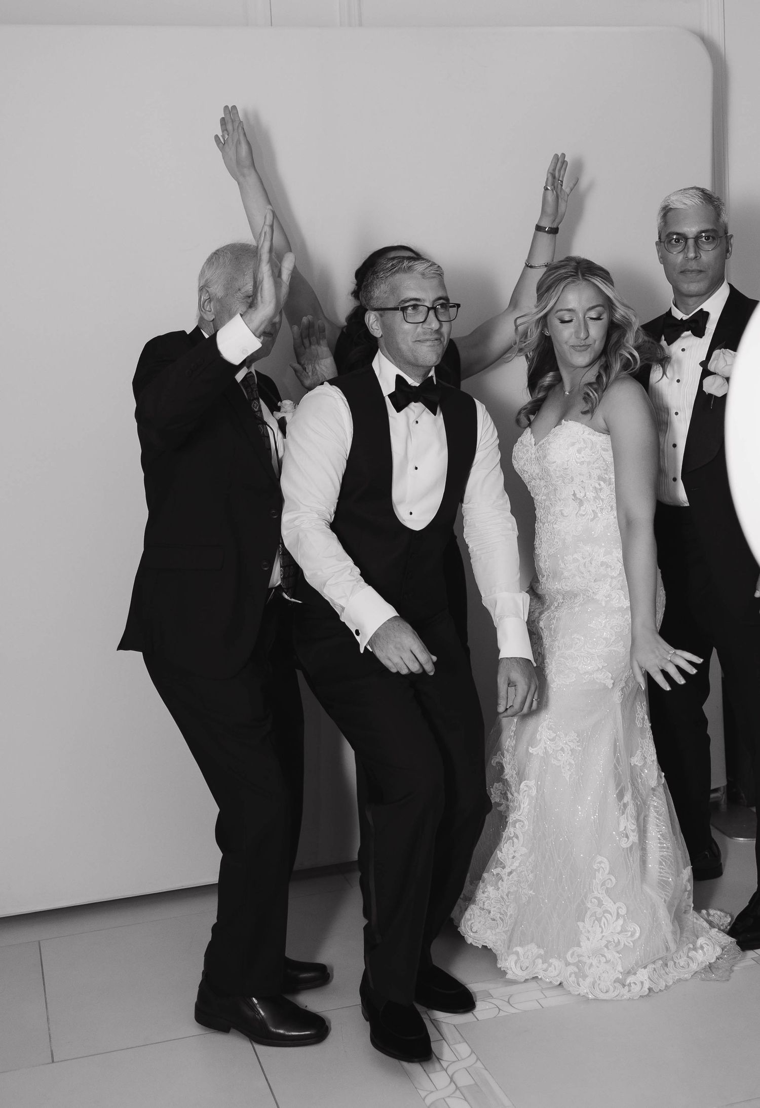 Wedding party poses playfully against white wall in formal attire.