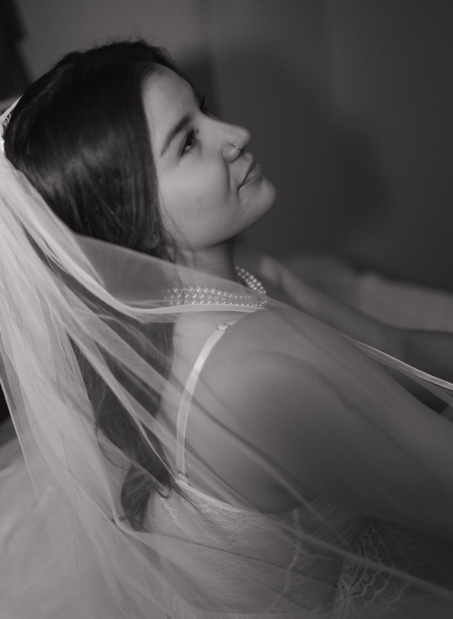 A black and white bridal portrait showing a delicate veil and pearl necklace detail in dramatic lighting.