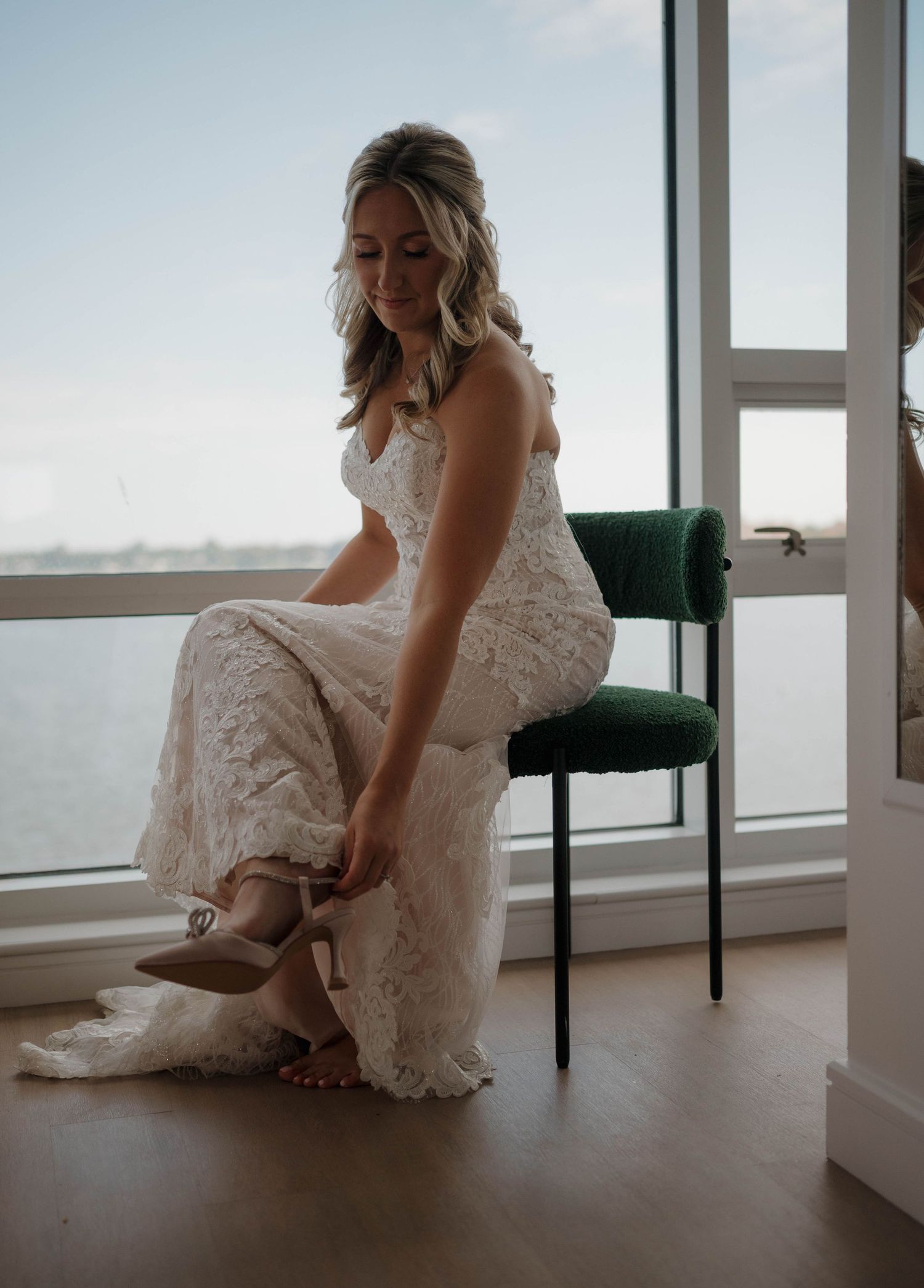 Bride in lace dress sitting by large windows with scenic view.