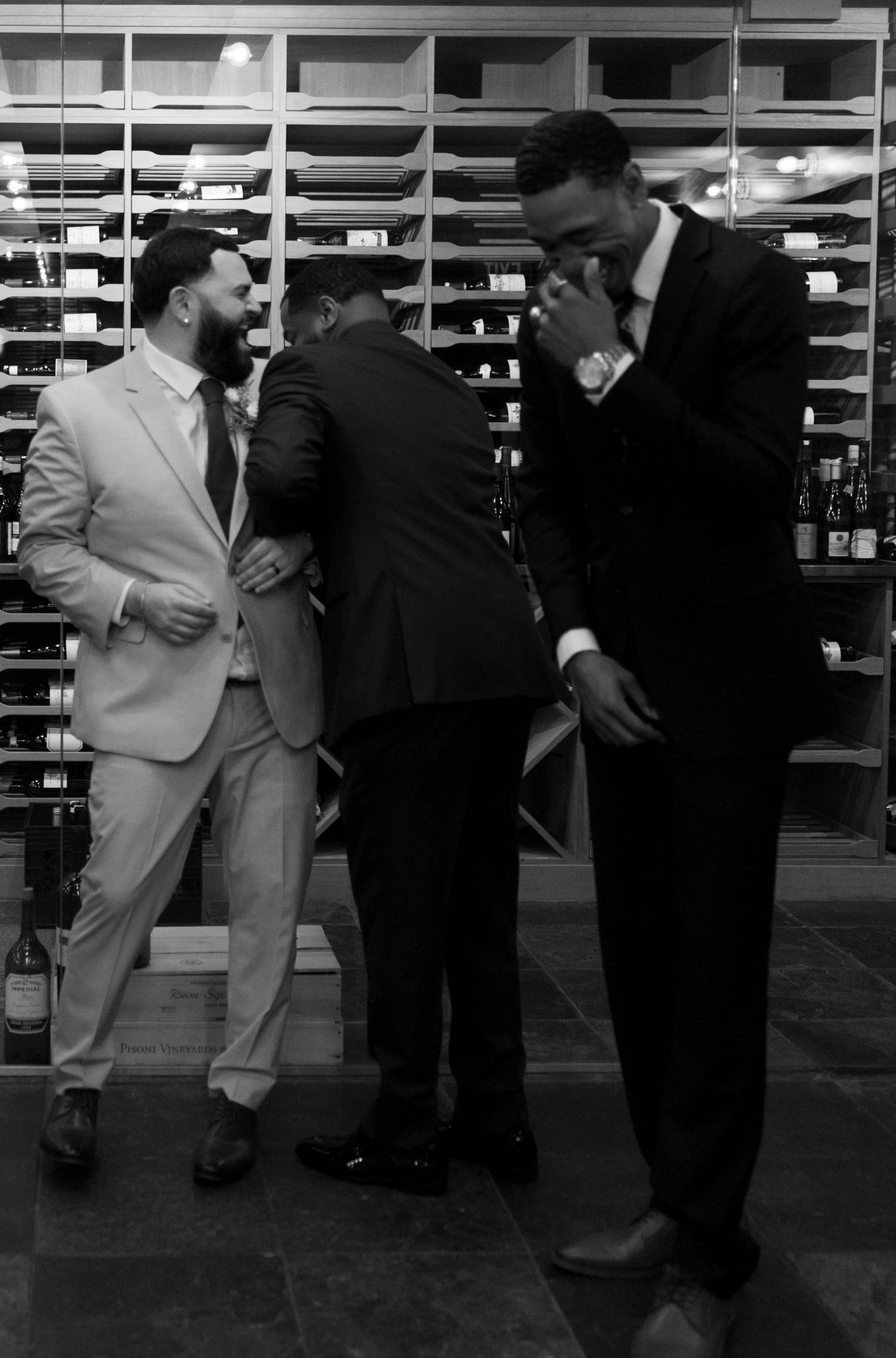 Wedding party members share laughs in a rustic wine cellar setting.