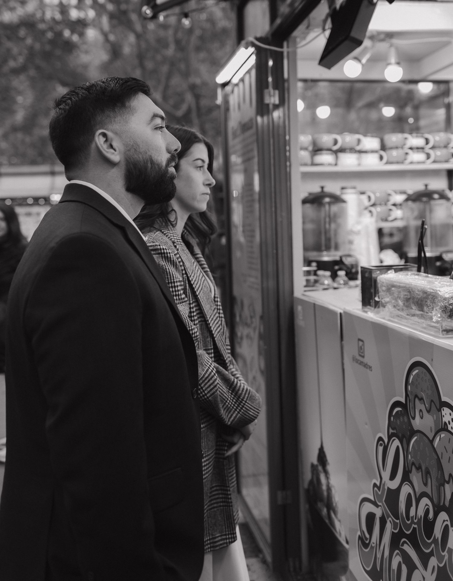 A couple gazes at a storefront display with glowing lights in a black and white photograph.