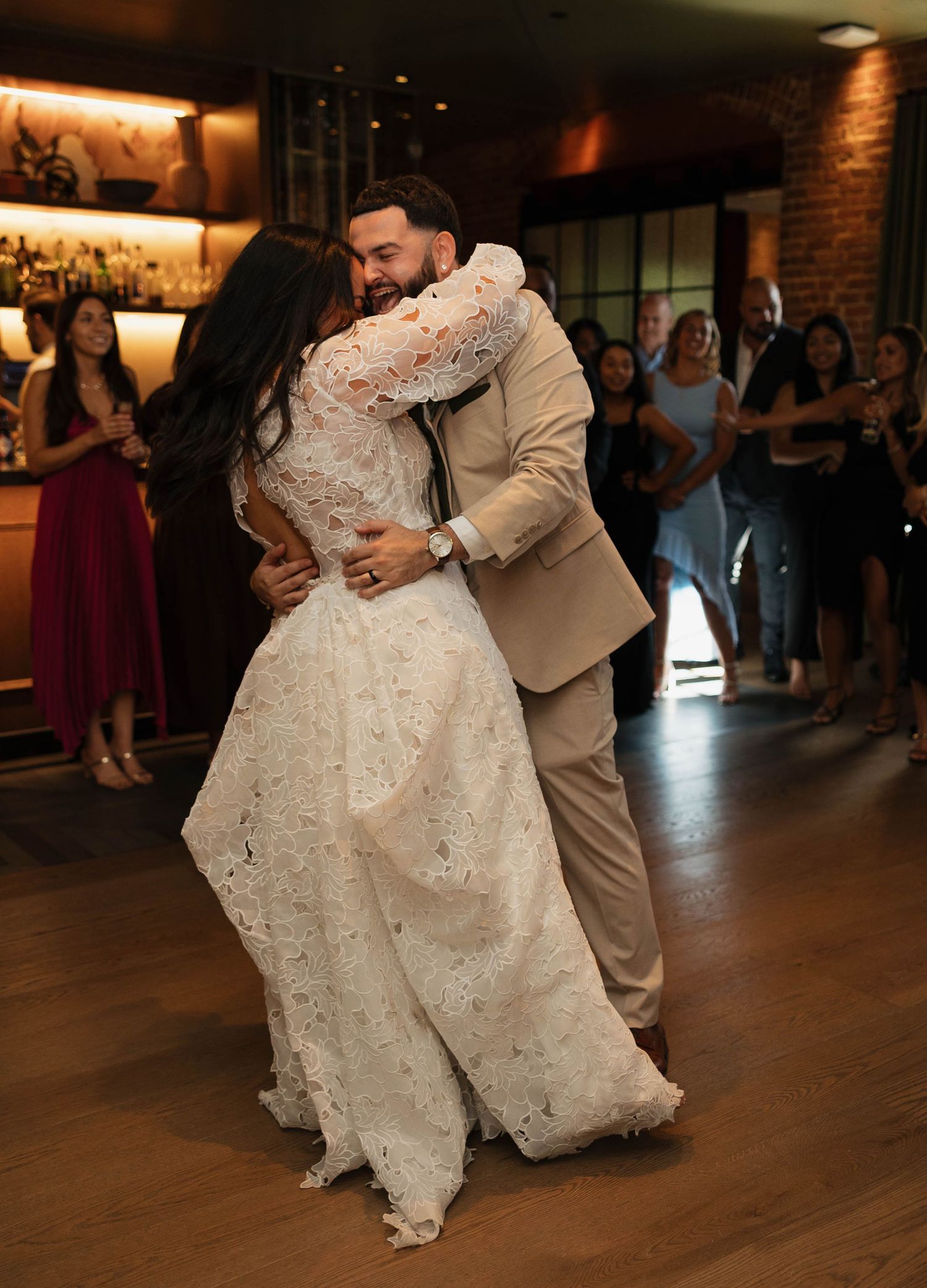 A couple dances together at a wedding reception, with the bride wearing a detailed lace dress and the groom in a tan suit.