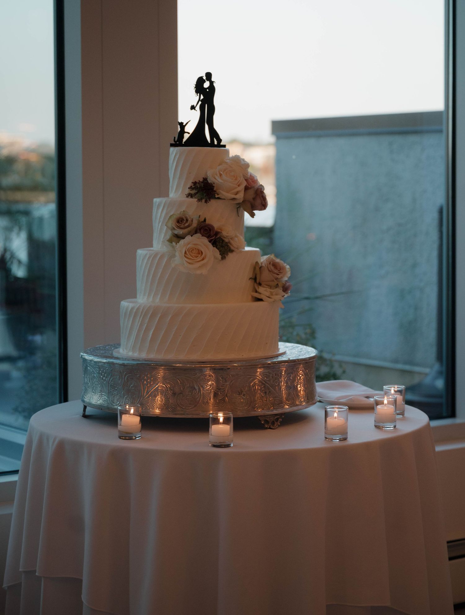 An elegant white wedding cake with floral decorations and a silhouette topper on a round table.