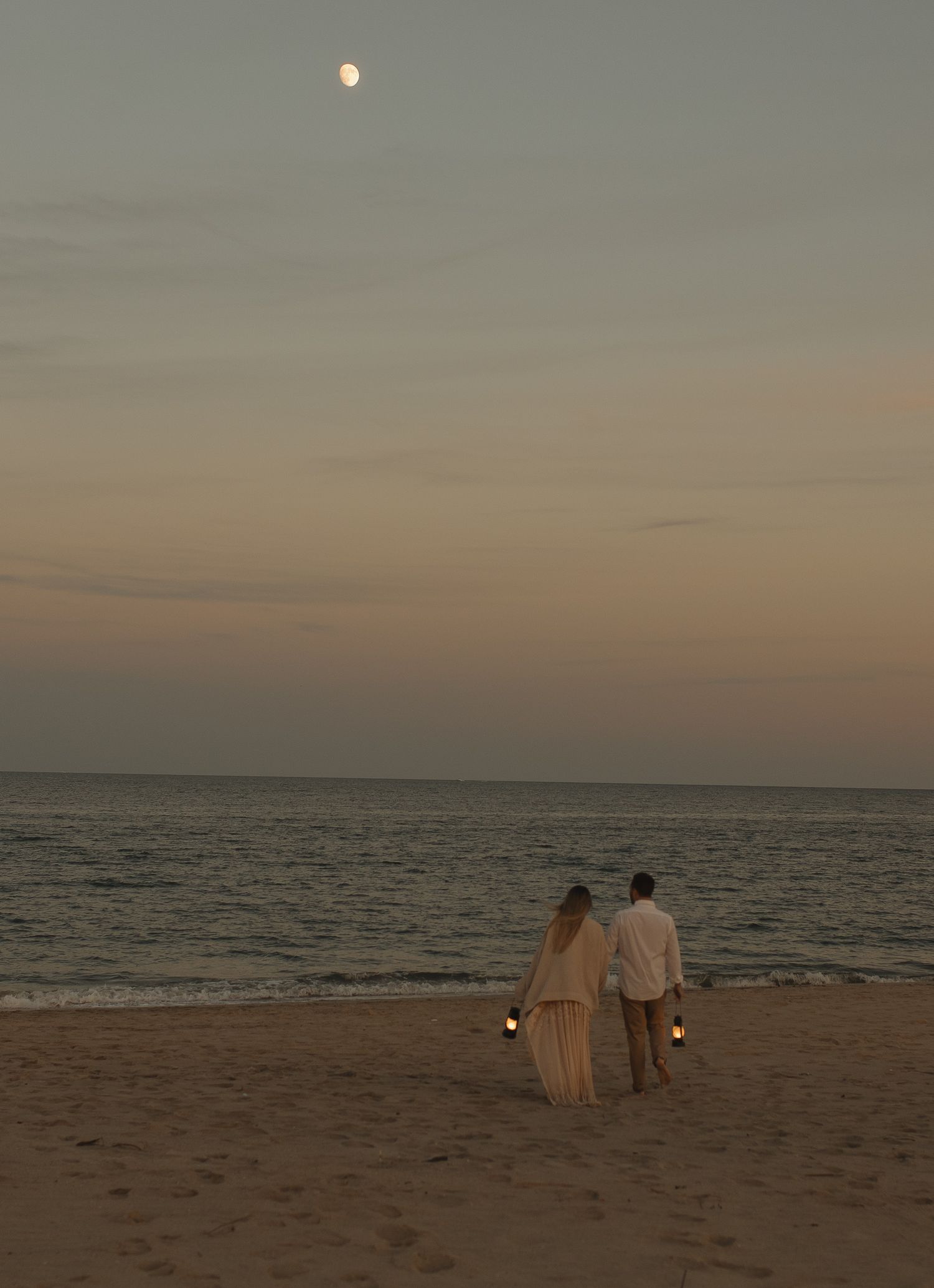 Two people in light-colored clothing walk along a serene beach at sunset with calm ocean waters in the background.