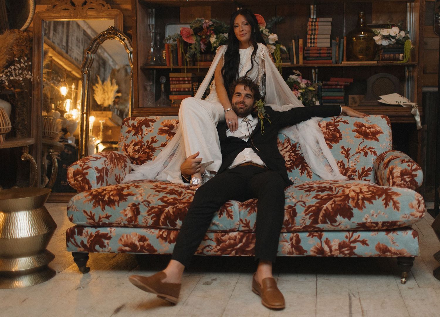 Three people pose casually on a floral print couch in a cozy vintage-styled room.