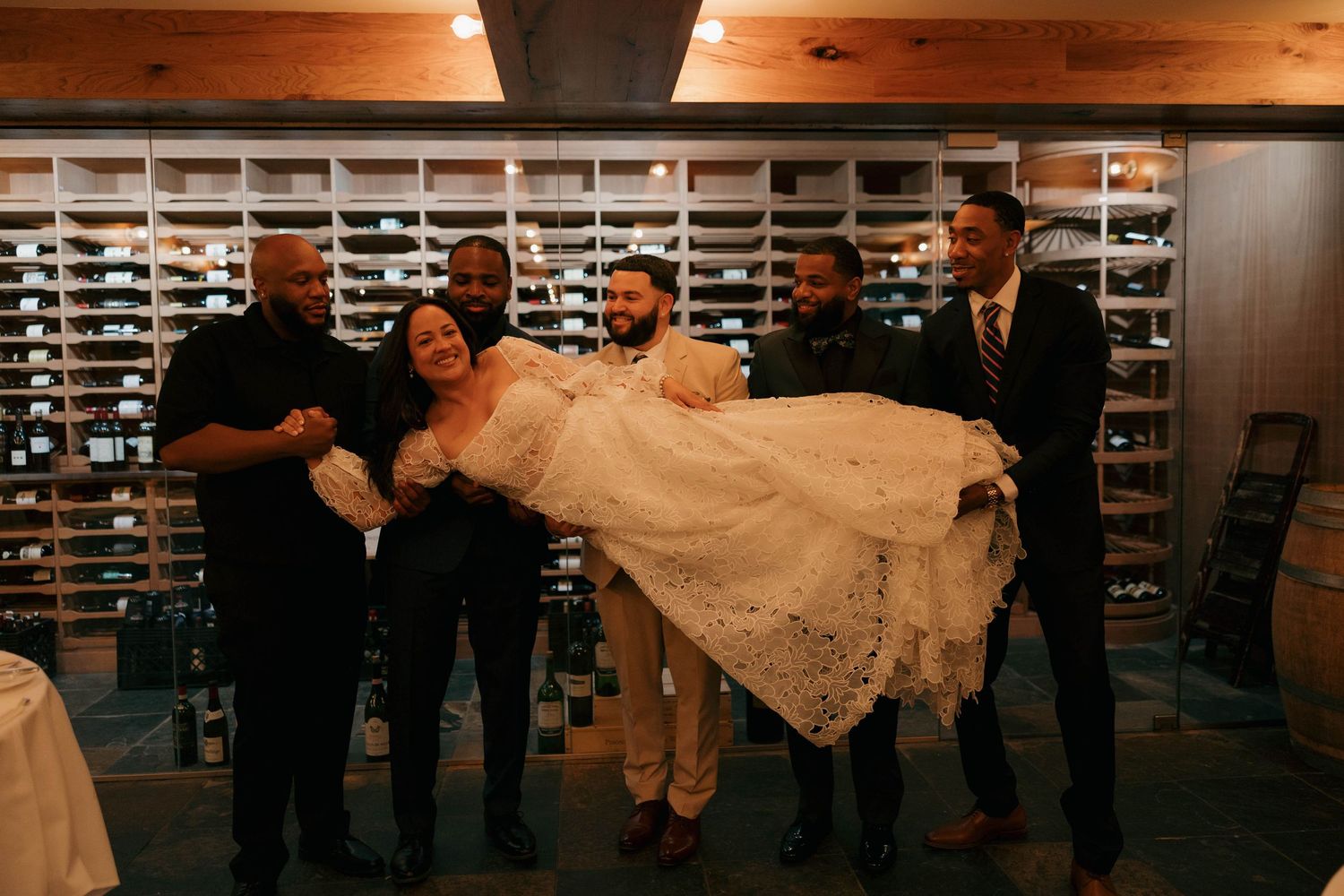Groomsmen playfully lift and carry someone in a wedding dress in wine cellar.
