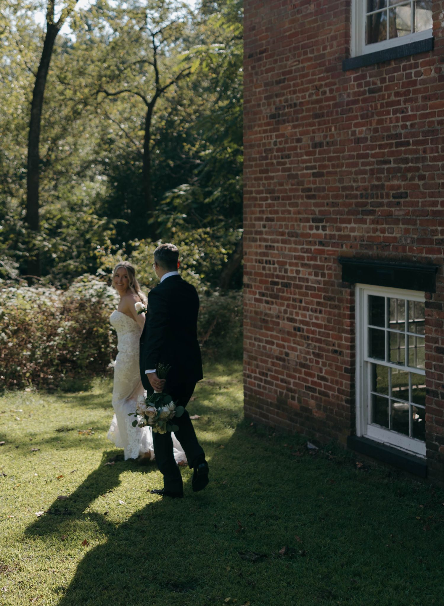 A couple walks along a brick building on their wedding day in a garden setting.
