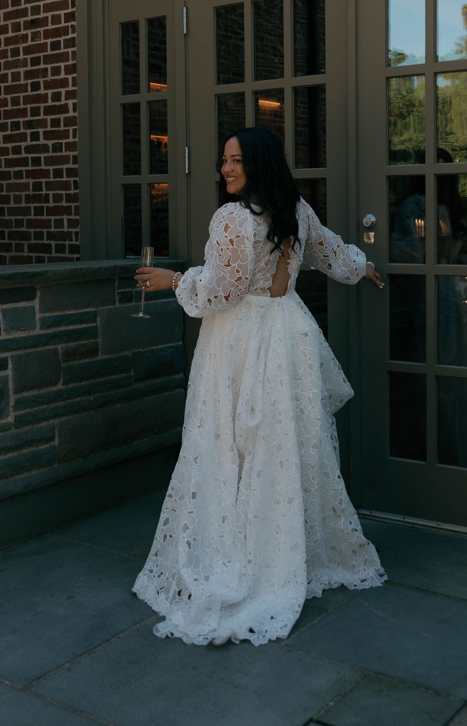 A bride in a flowing lace wedding dress poses with champagne against a brick building.