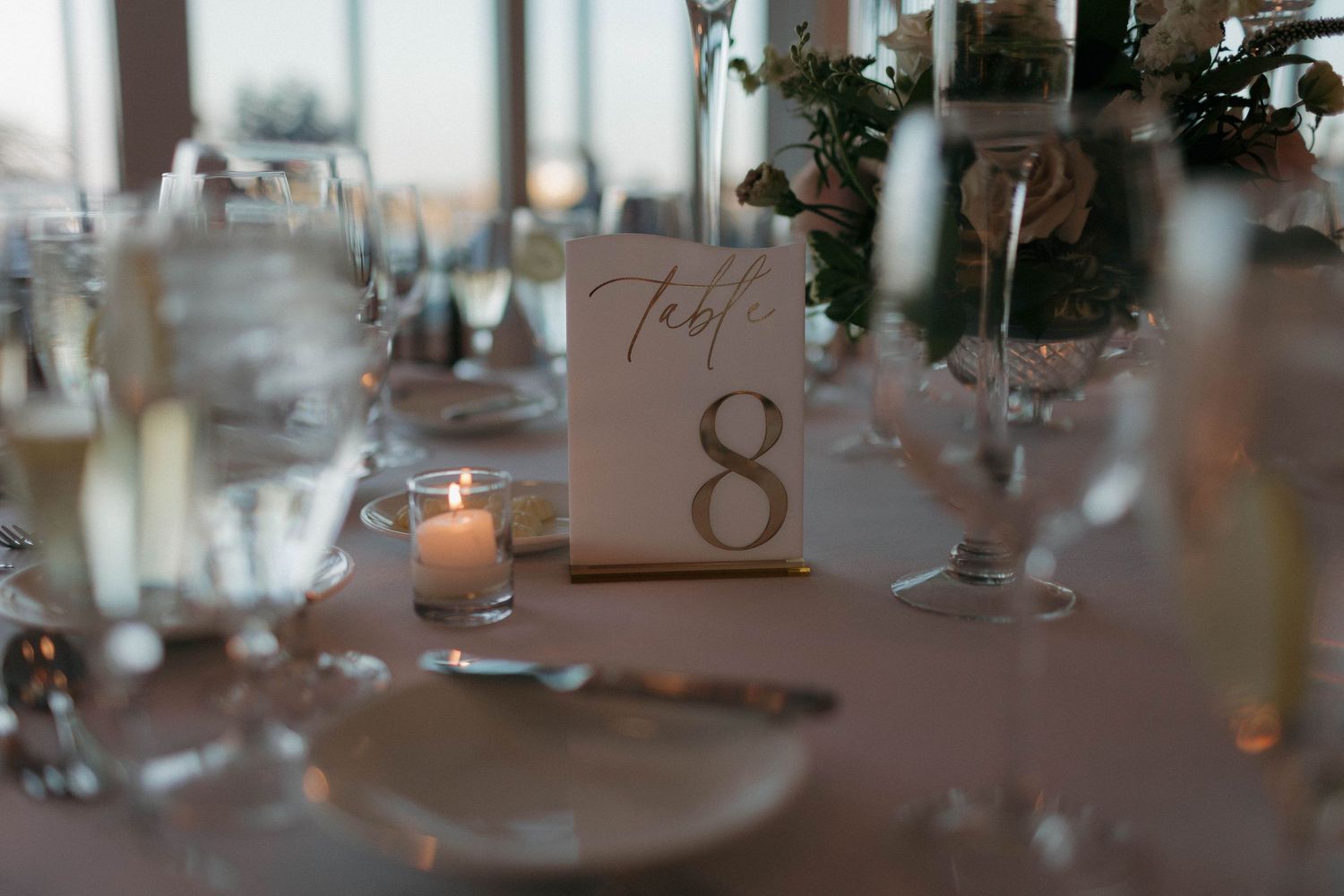 Table setting with number 8 card holder, glassware and candlelight at wedding reception.