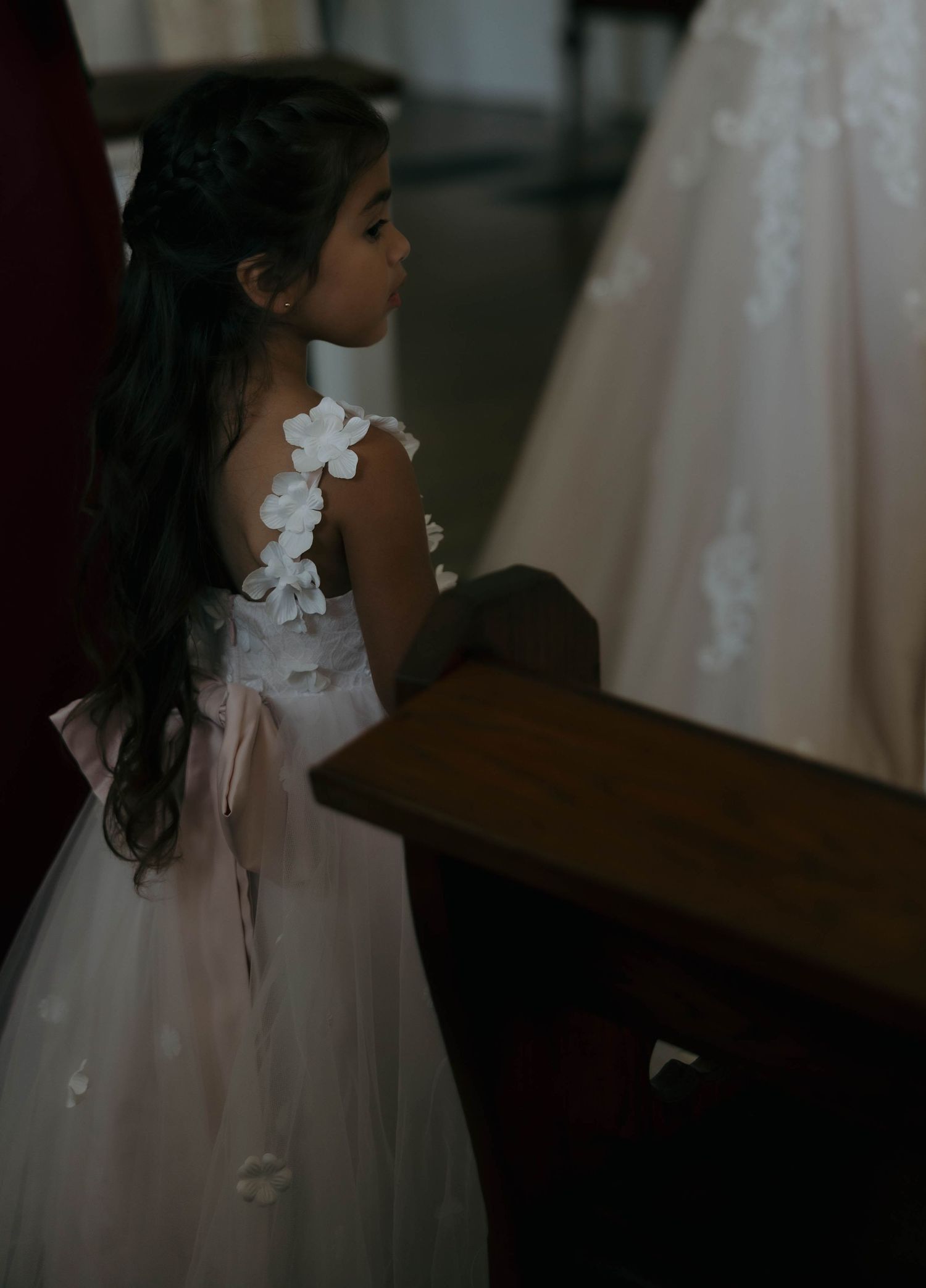 A flower girl in a delicate white dress with floral details stands in soft lighting.