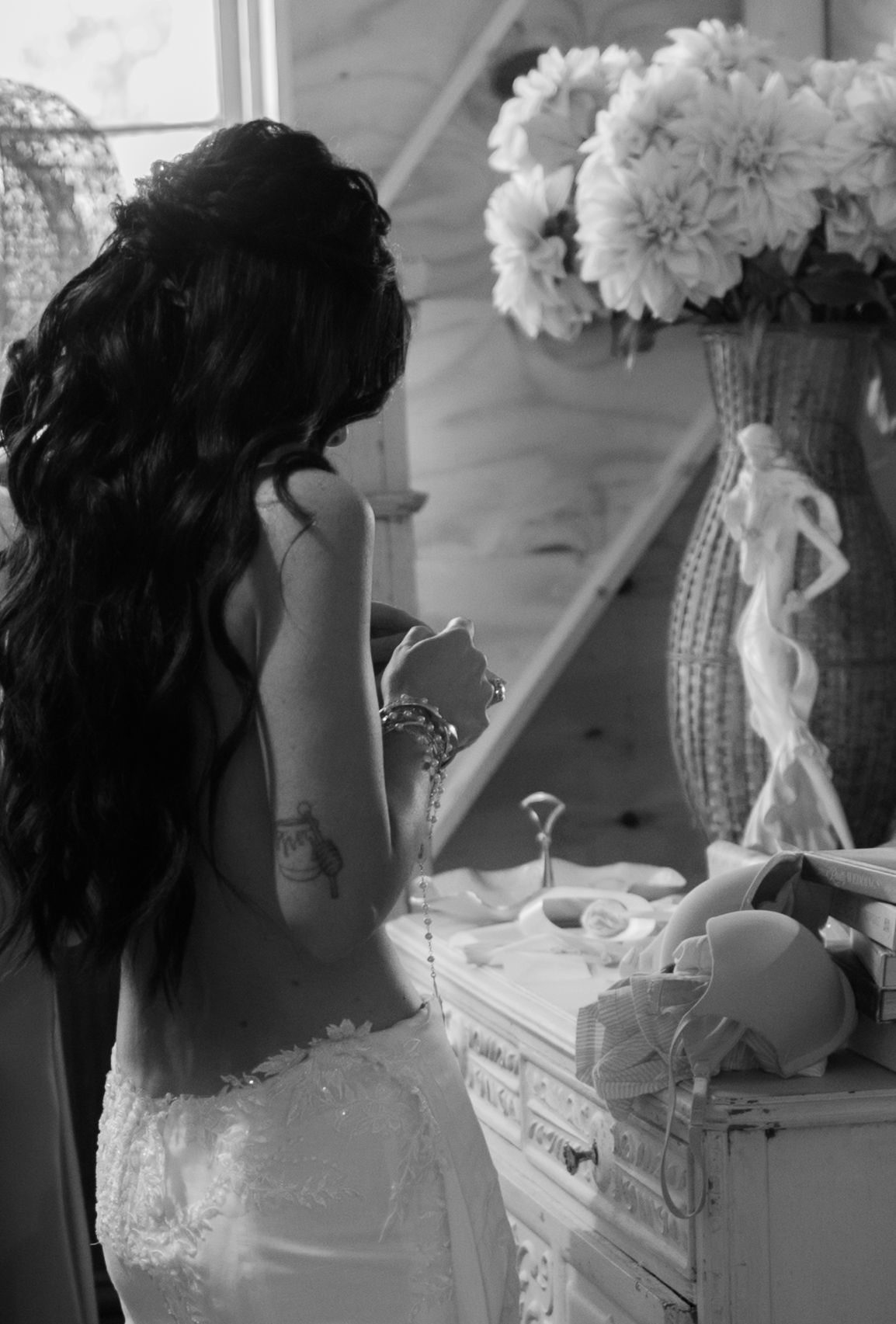 A woman in a wedding dress with long dark hair stands at a vanity with flowers and decorative items.