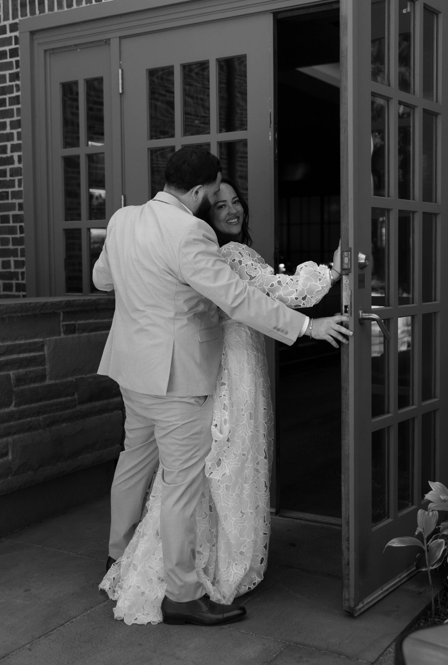 A tender black and white wedding photo showing a couple embracing by glass doors.