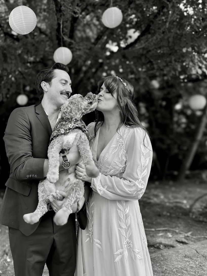 Black and white photo of a wedding couple holding their dog, with the bride kissing the dog and lanterns in a tree