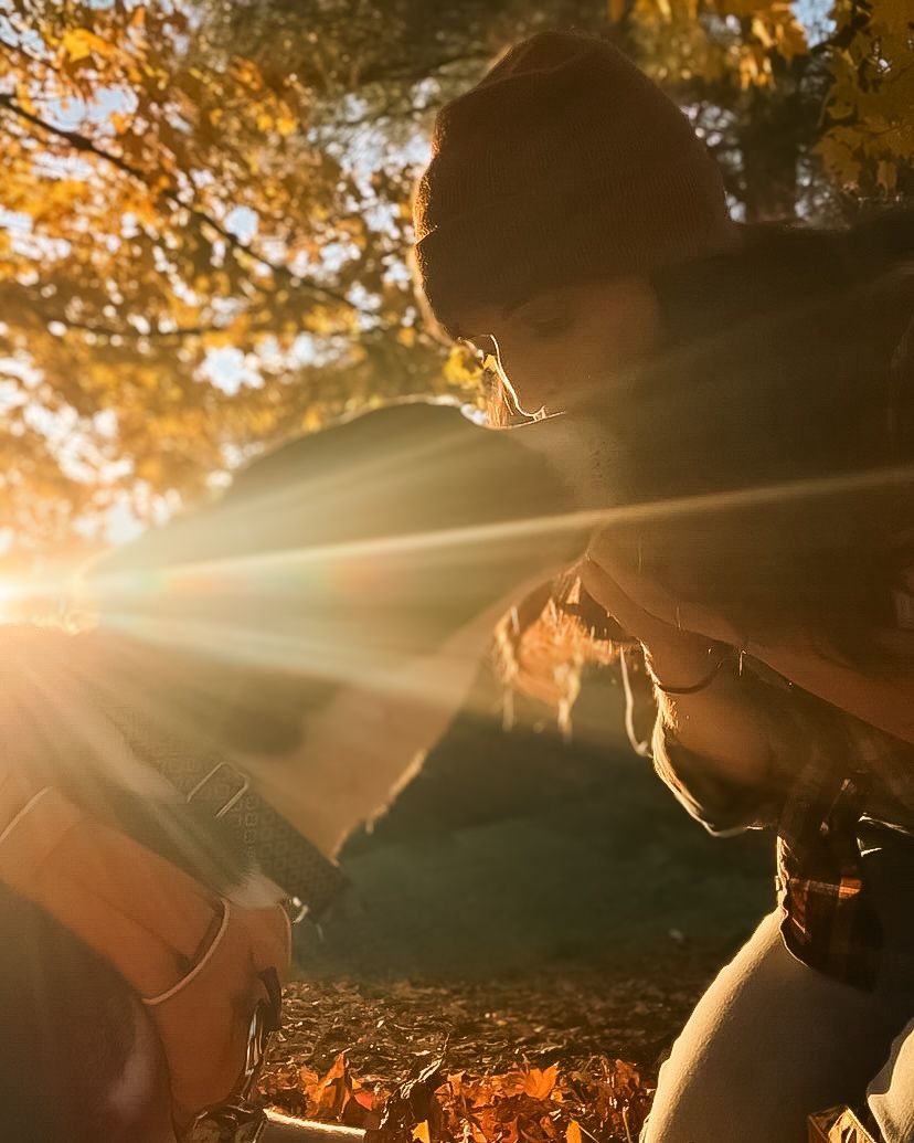 Woman in a hat kissing her dog on the forehead at sunset, with fall foliage, scattered leaves, and a warm sunburst