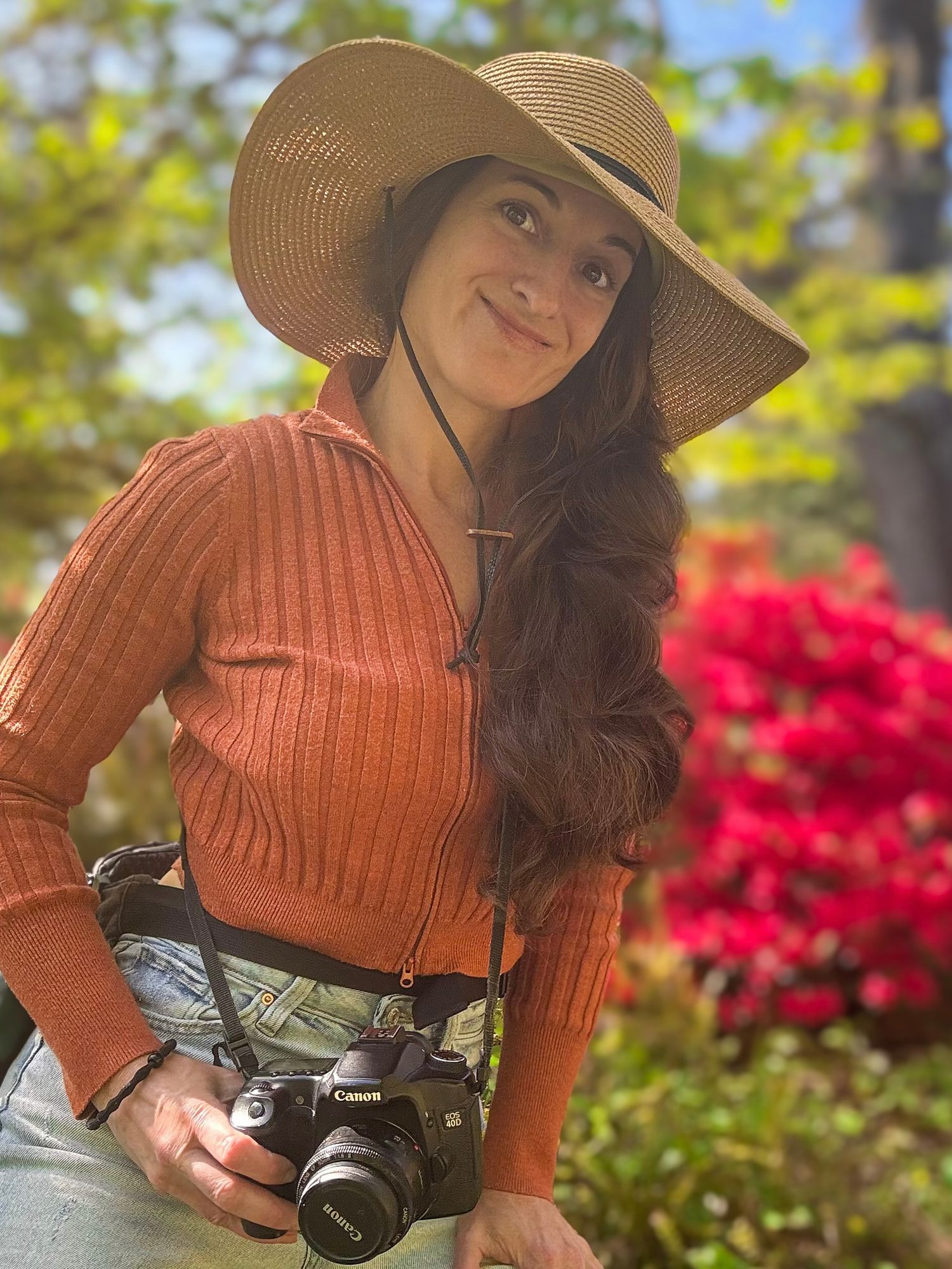 Woman wearing a hat and orange sweater holding a camera, standing near a blooming azalea bush under blue sky.