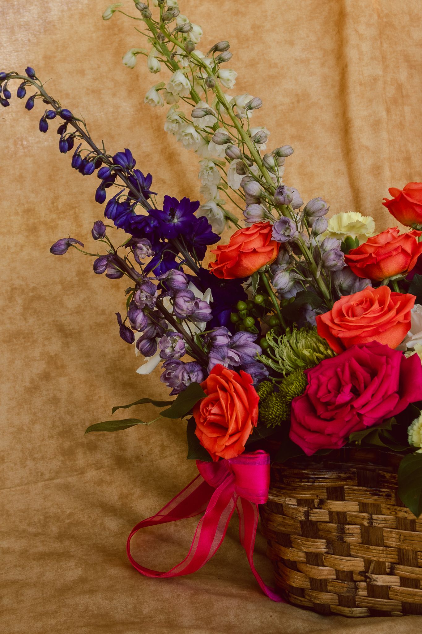 Colorful floral arrangement in woven basket with red roses, purple delphiniums, and pink ribbon on golden background.