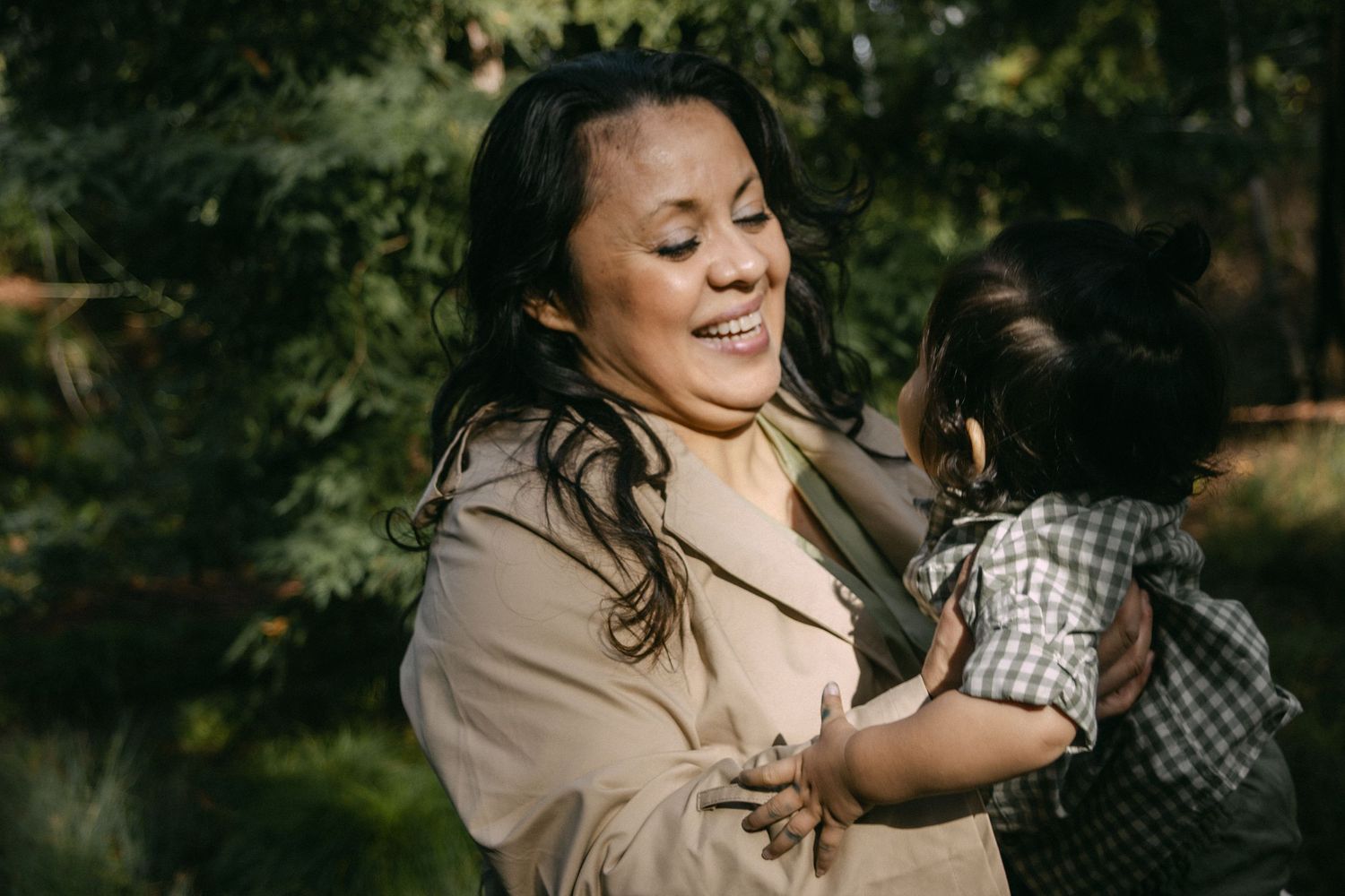 A person wearing a beige jacket laughs joyfully while holding a small dog outdoors in natural lighting.