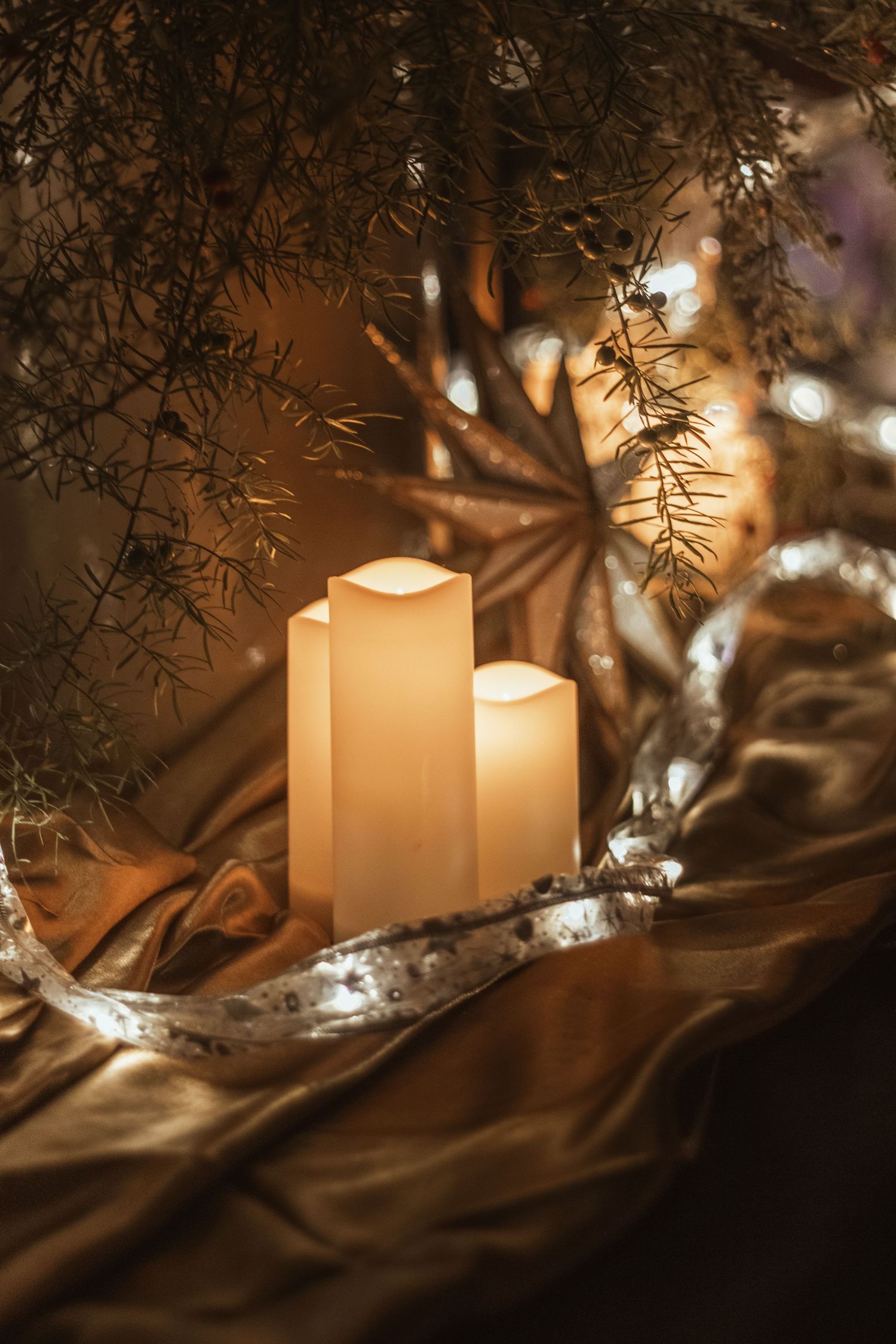 Glowing white pillar candles nestled under pine branches on golden satin fabric with silver star decorations.