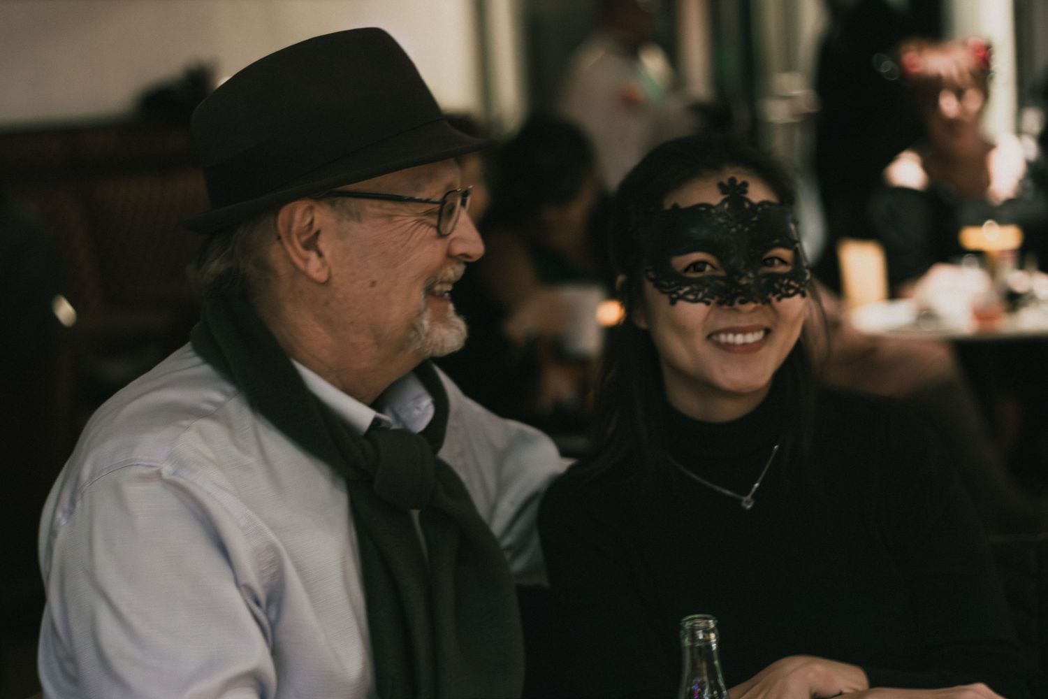 An older man in a hat and a woman wearing a decorative masquerade mask smile together at a dimly lit event.