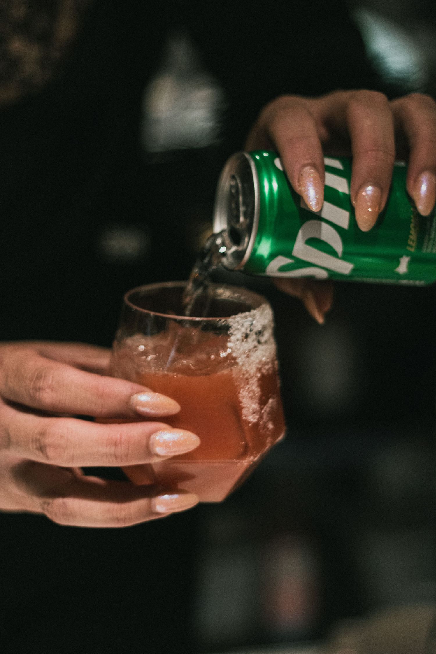Hands pouring a carbonated beverage into a dark cocktail glass creating a foamy effect in low light.