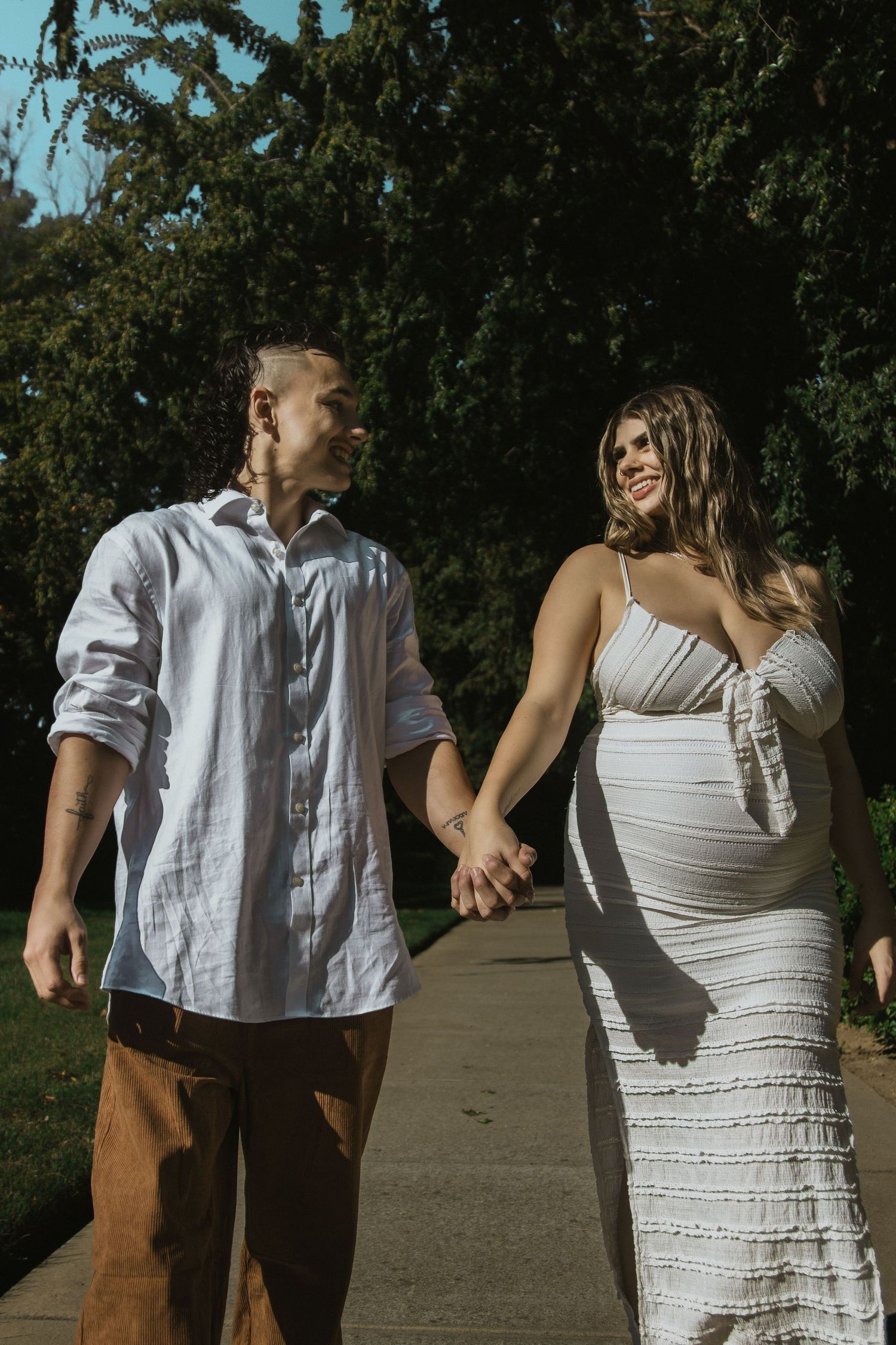 Couple in white clothing walking hand in hand on a concrete path outdoors.