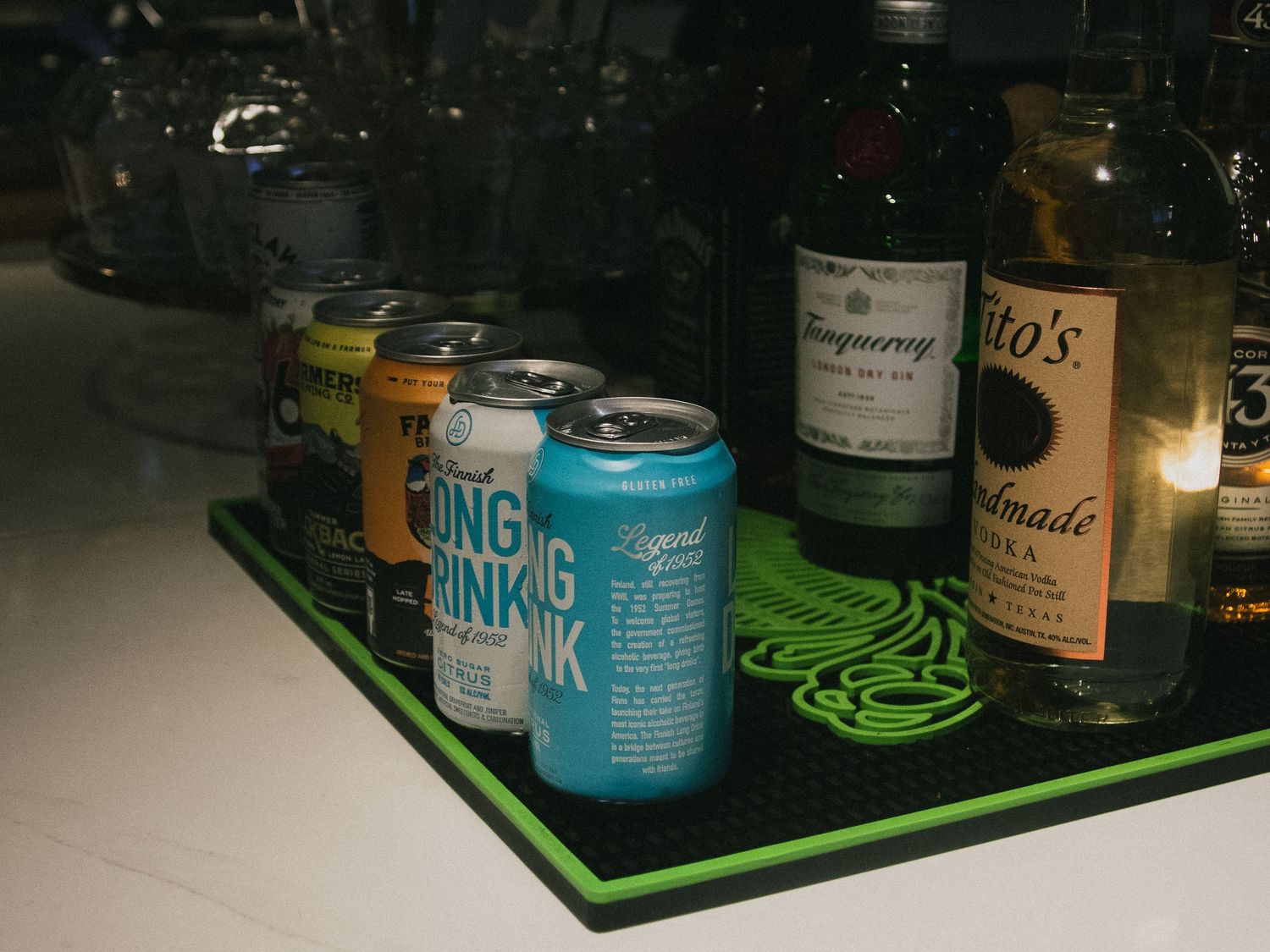 Assorted beverage cans and liquor bottles including Tanqueray gin and Tito's vodka displayed on a green bar mat.