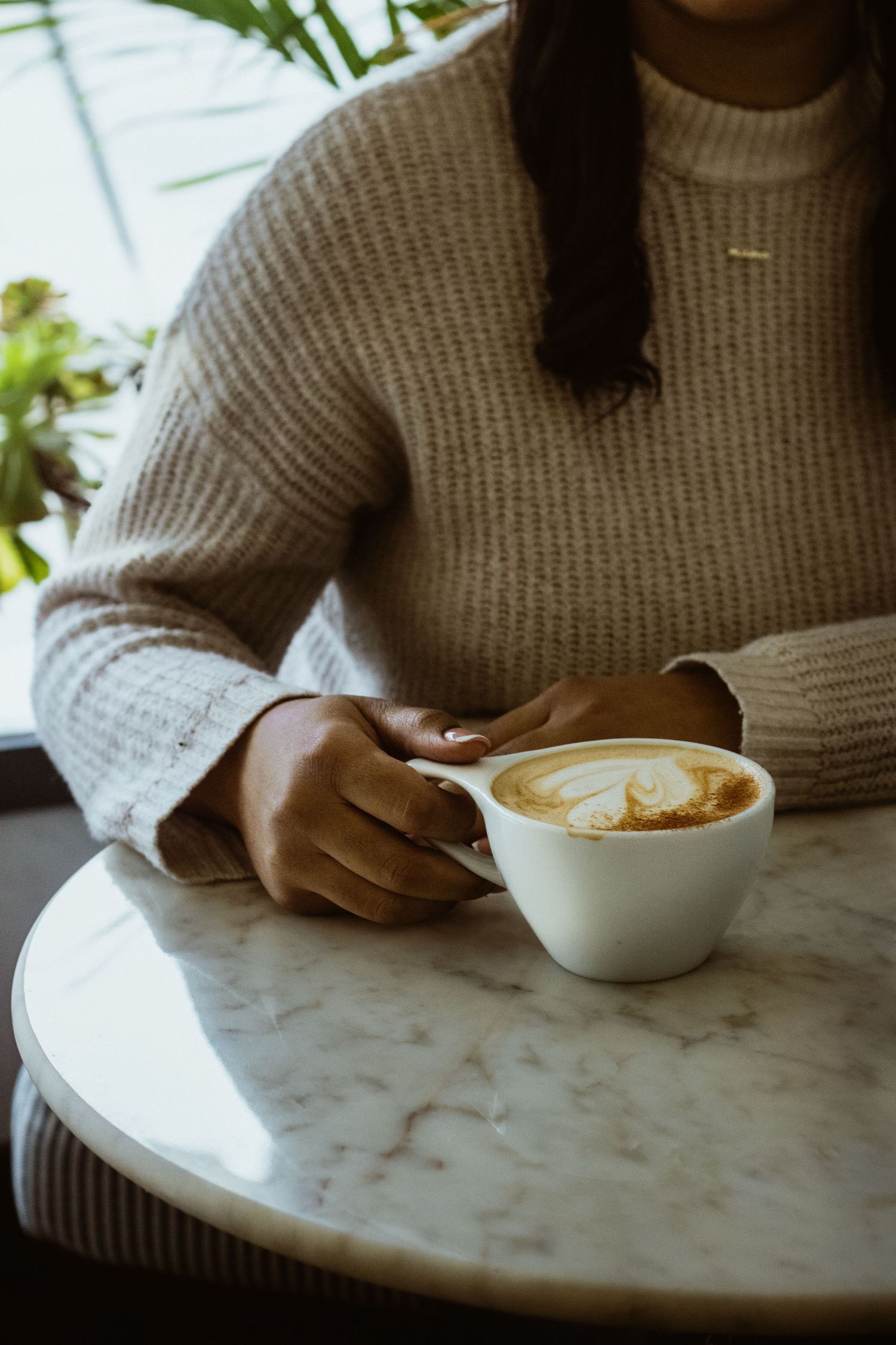 Someone in a cozy beige sweater enjoys a latte with latte art on a marble table at a cafe.