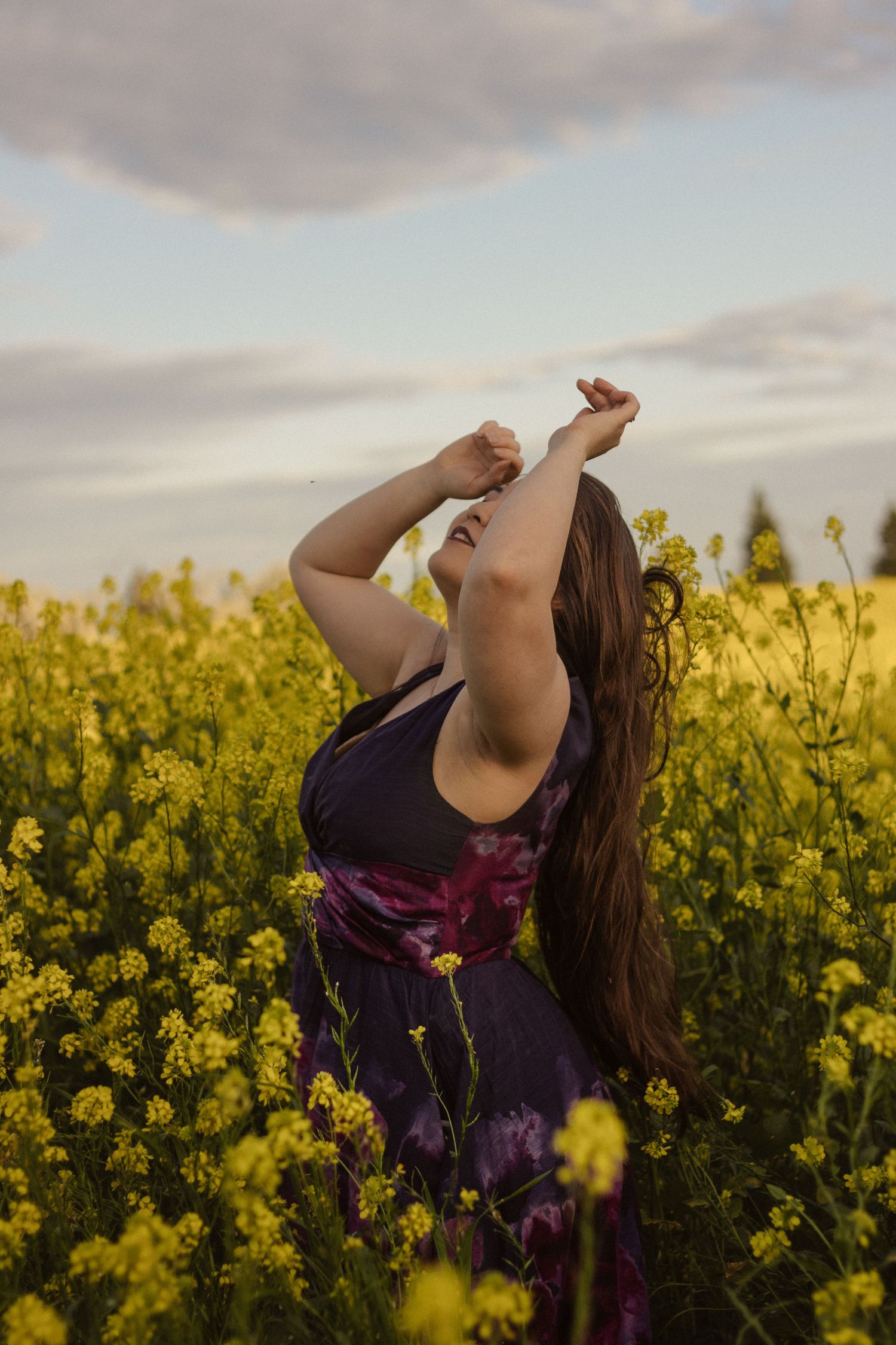 Dramatic pose in purple dress surrounded by bright yellow canola flowers during golden hour.