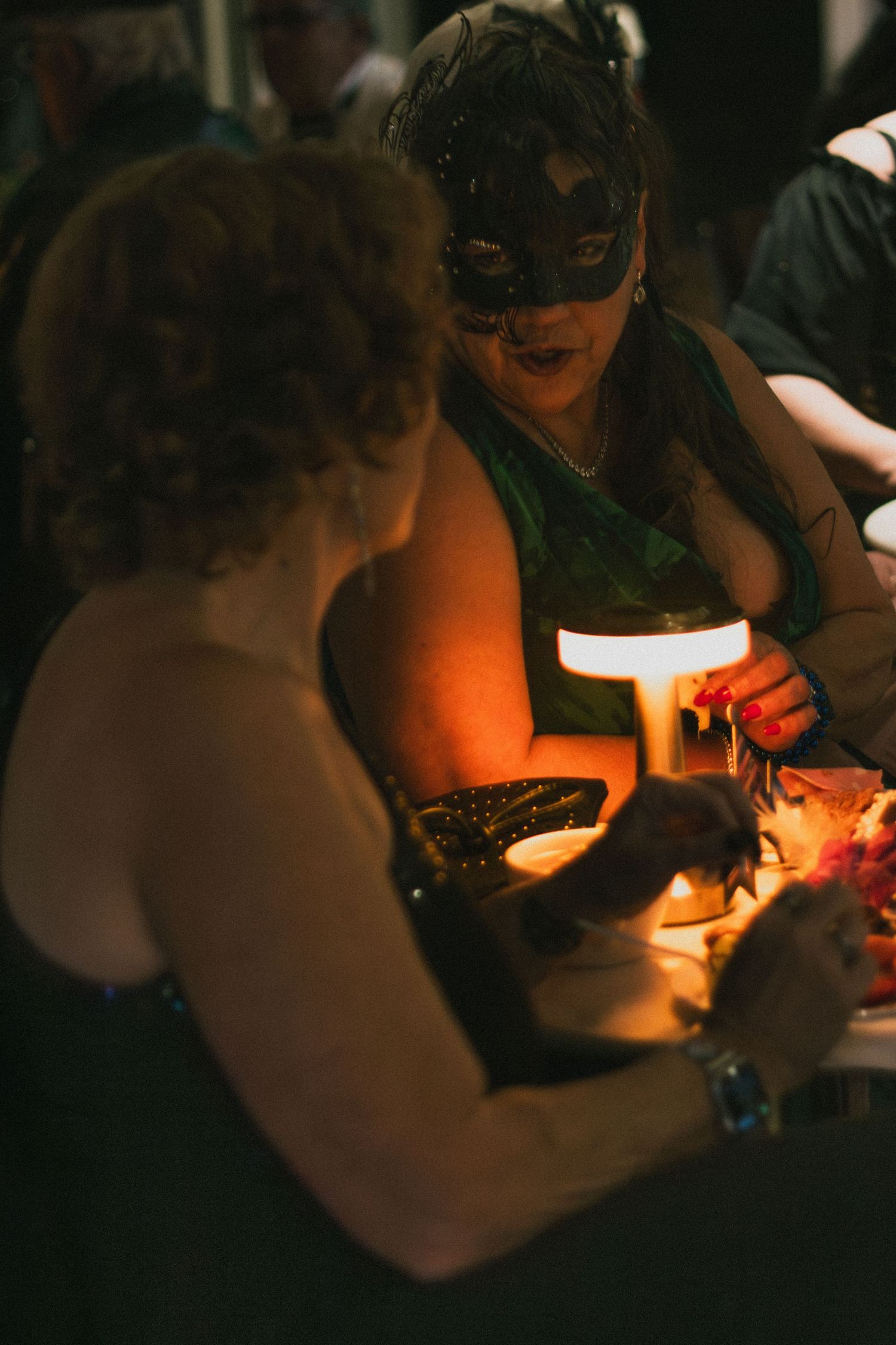 Two women at a masquerade party, one wearing a black mask, gathered around a glowing lamp at a dimly lit table.