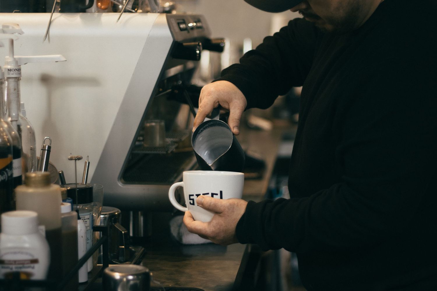 A person pouring steamed milk into a coffee cup creating latte art in a dimly lit coffee shop setting.