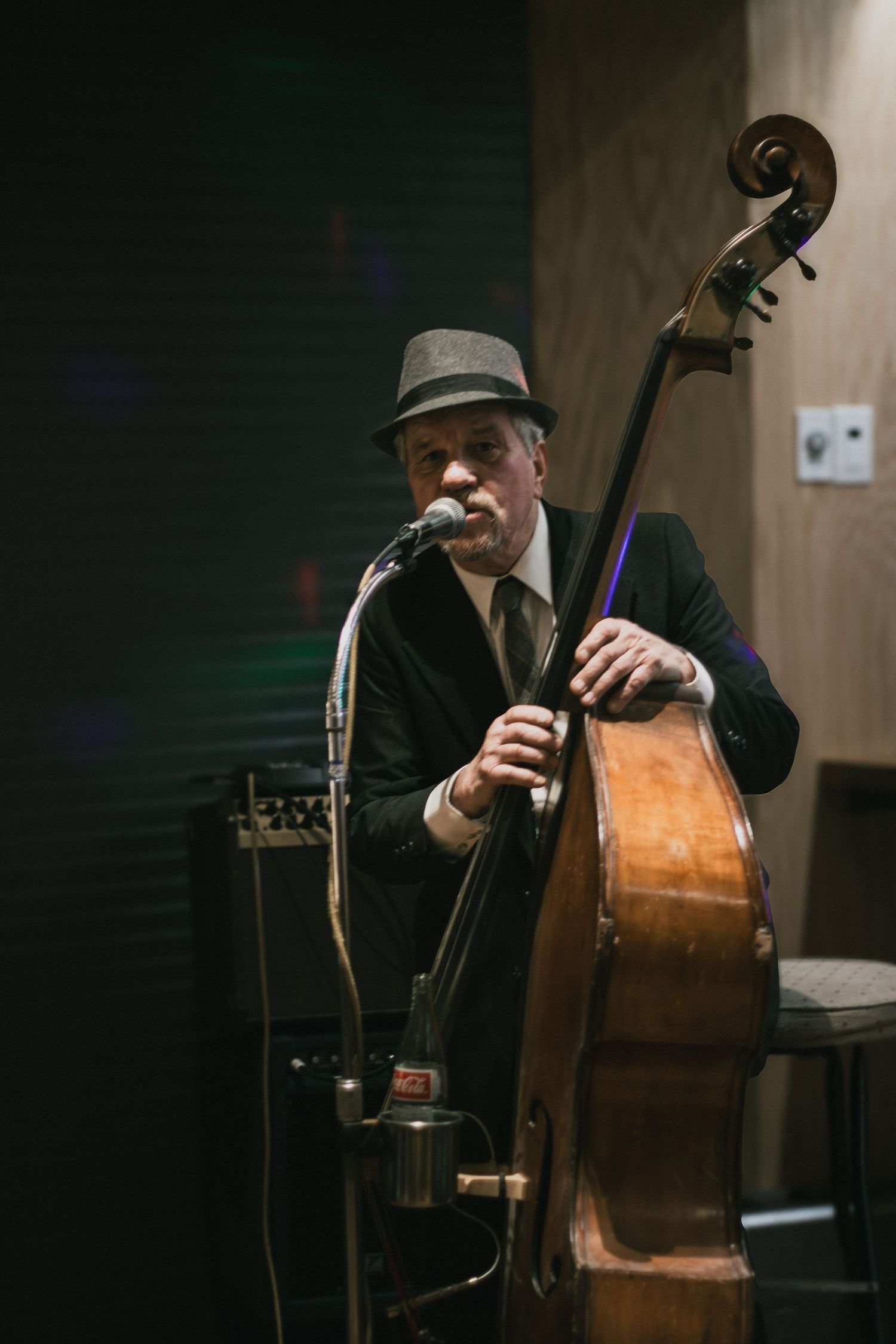 A jazz musician in a fedora performs with an upright bass and microphone in a dimly lit venue.