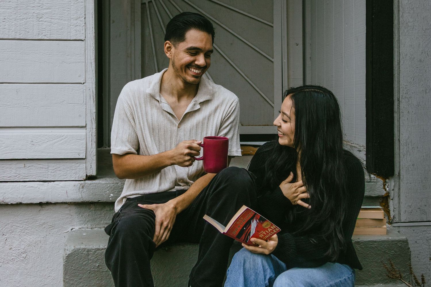 Two people relaxing on porch steps with books and beverages.