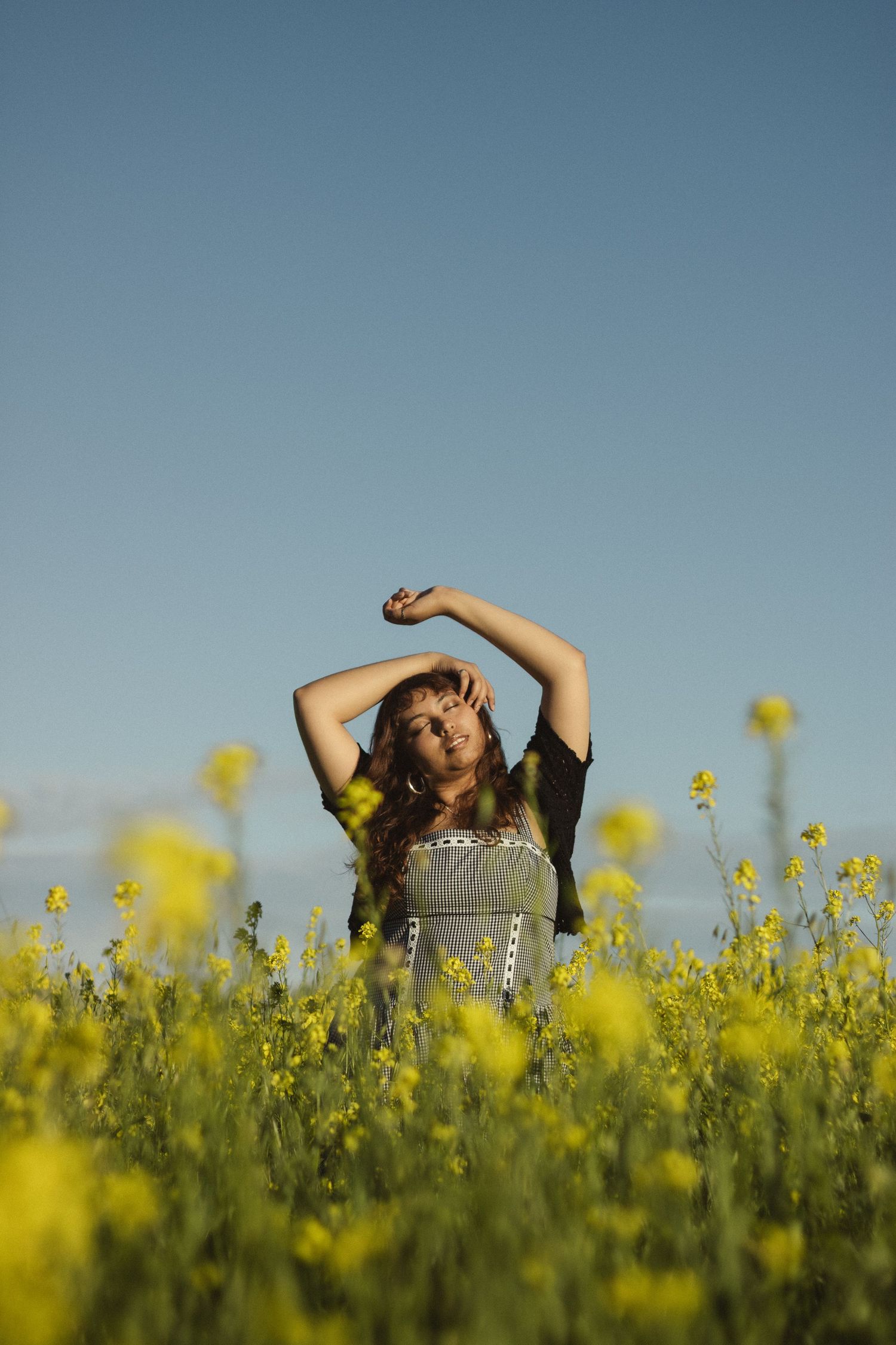 Person lying in vibrant yellow canola field under blue sky with arms raised gracefully.