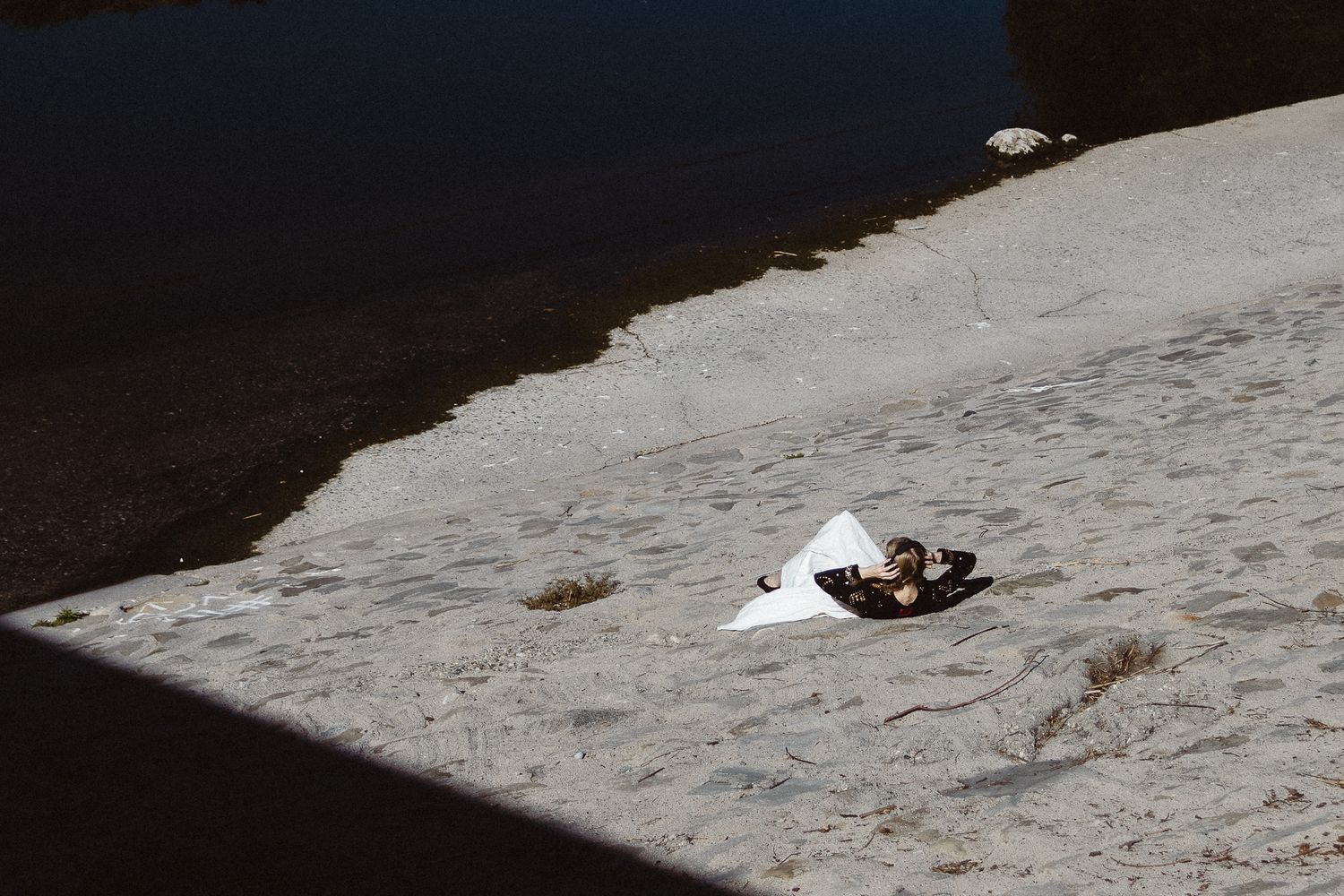 Litter and debris scattered on a sandy riverbank beside calm dark water in natural outdoor setting.