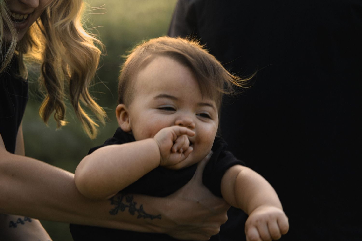 A joyful toddler in a black t-shirt laughs with their hand near their mouth while being held up in golden sunlight.