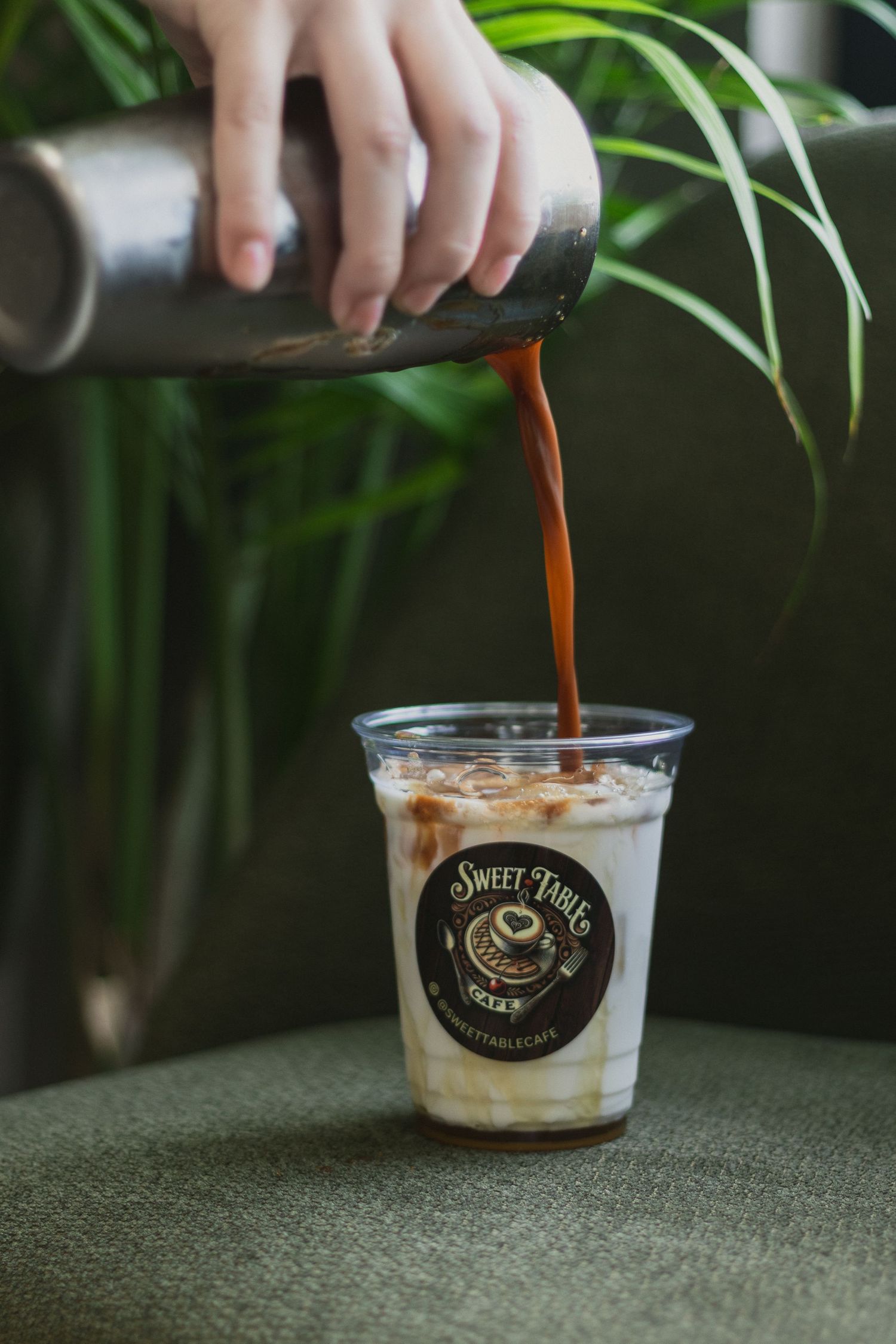 Dark coffee being poured from a metal container into a clear plastic cup with Sweet Time's cafe branding on green fabric.