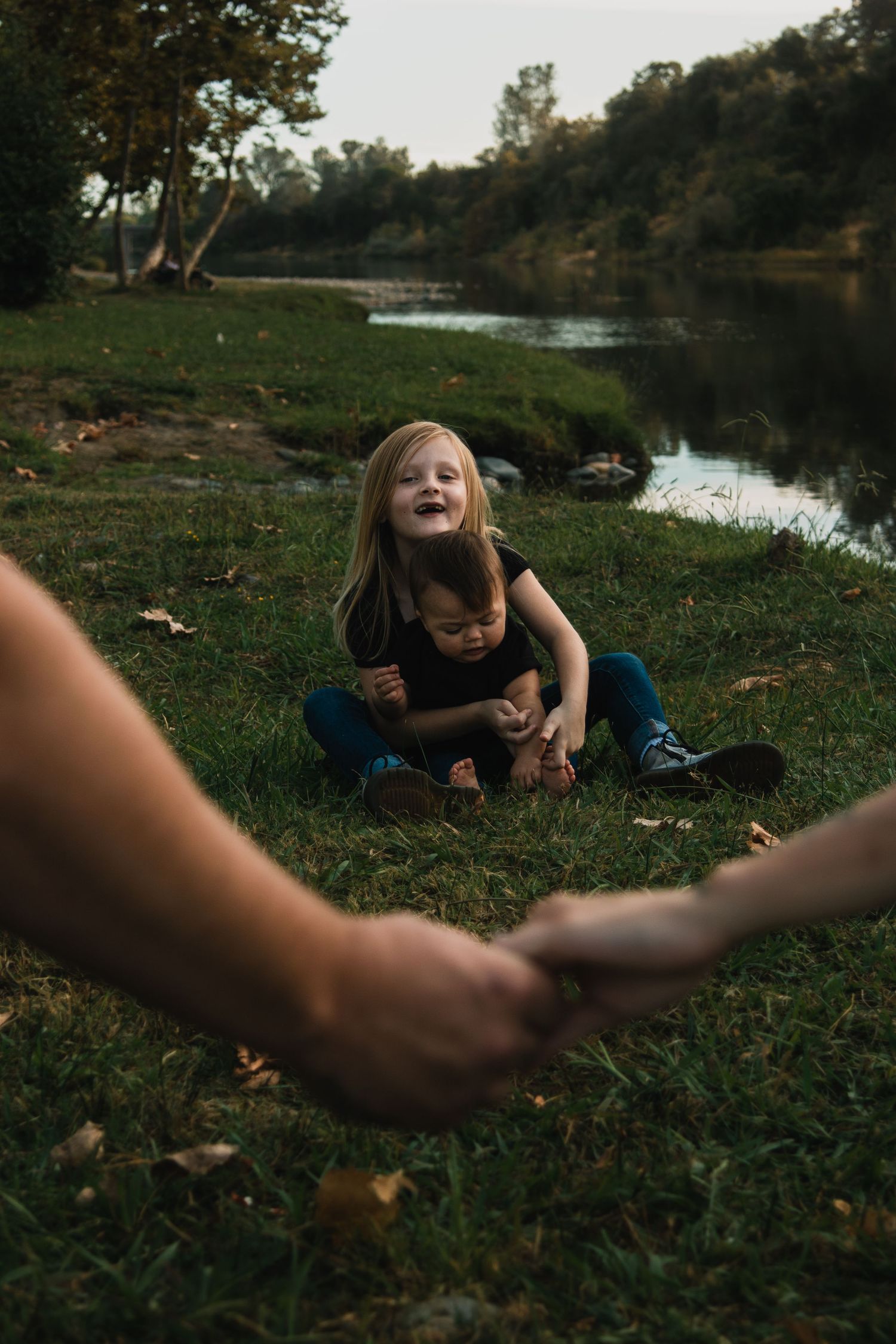 A playful scene on grassy riverbank with arms reaching towards someone lying on the ground.