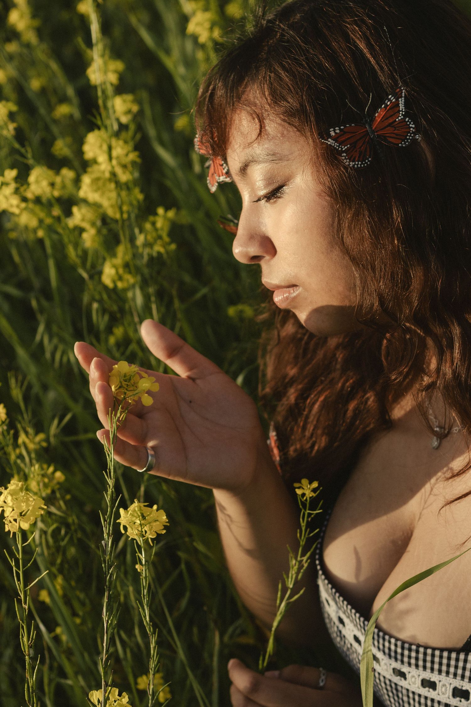 A person in gingham dress gently touches yellow wildflowers in golden sunlight during a warm summer evening.