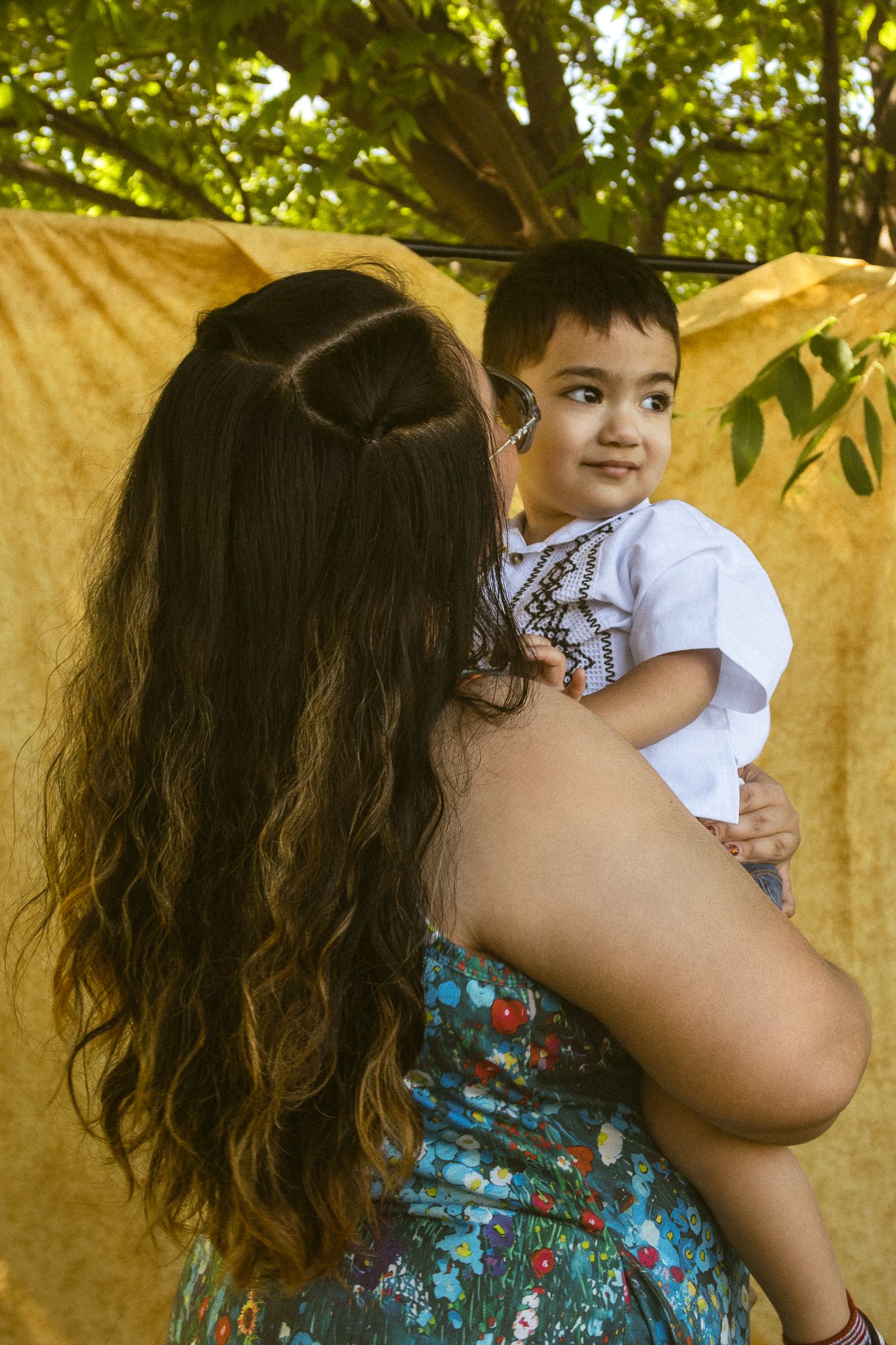 A woman with long curly hair holds a smiling baby in a white shirt outdoors near a yellow backdrop.