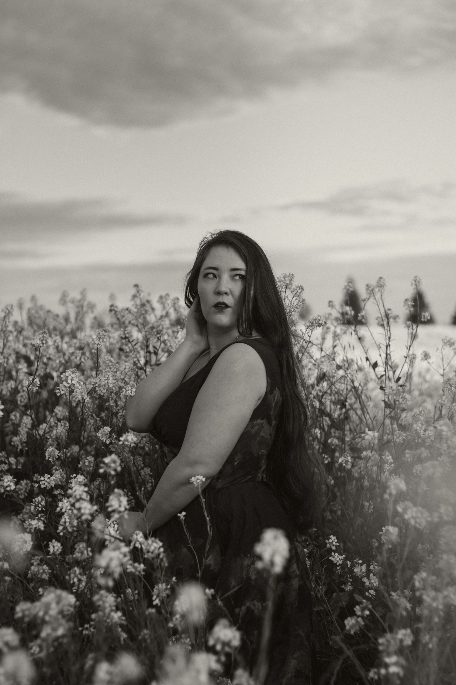 A dreamy black and white portrait shows a person among blooming wildflowers against a moody sky.