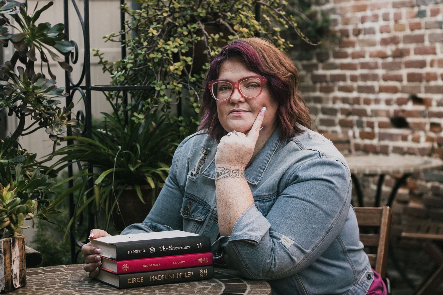 Person in glasses sitting at brick wall with plants and books in background.