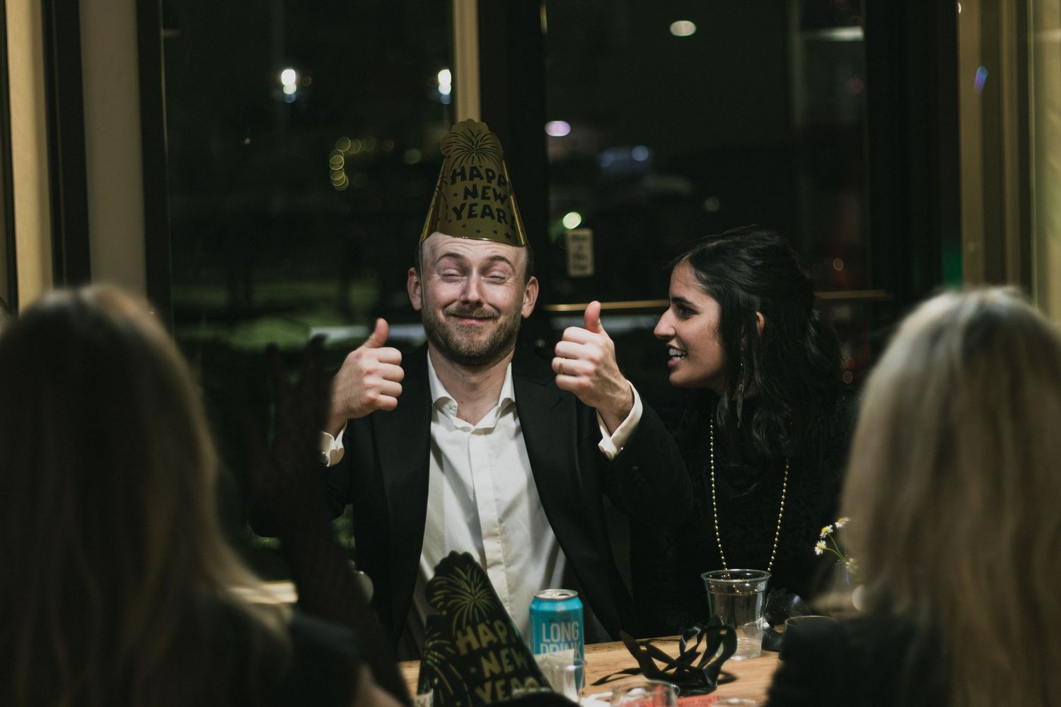 Man in party hat giving thumbs up at New Year's Eve celebration with smiling woman at restaurant table.