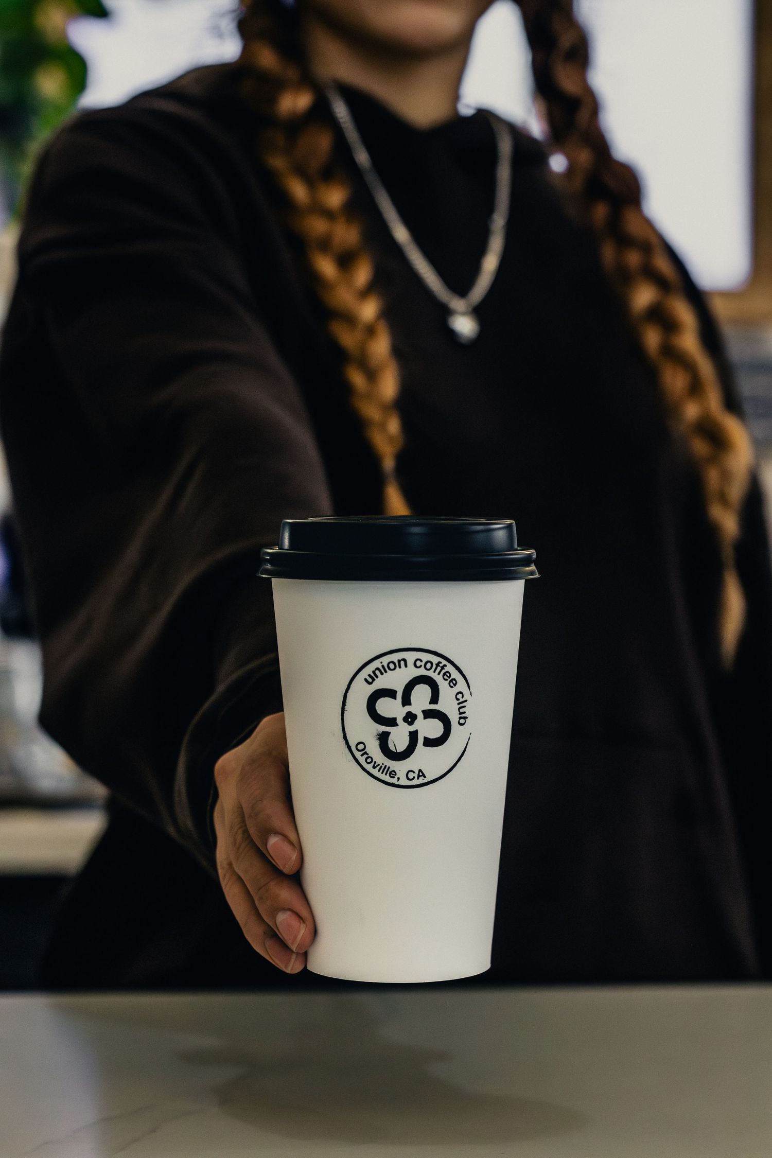 A person in black holds a white coffee cup with a circular logo while wearing braided hair and a necklace.
