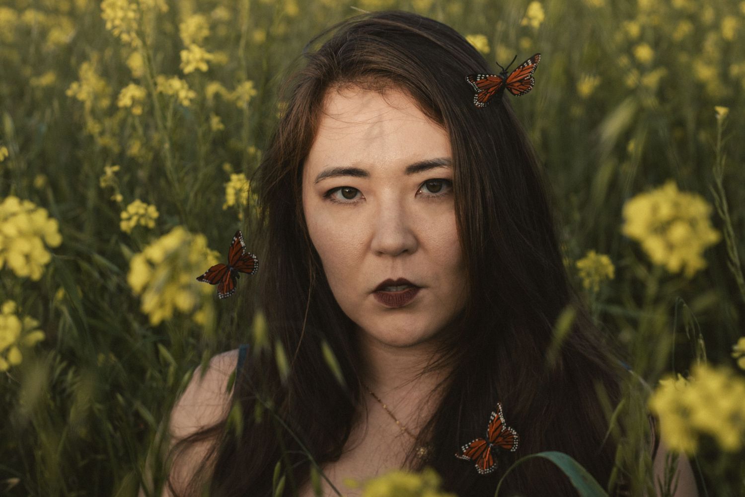 Portrait in yellow canola field with monarch butterflies and dark makeup creating ethereal mood.