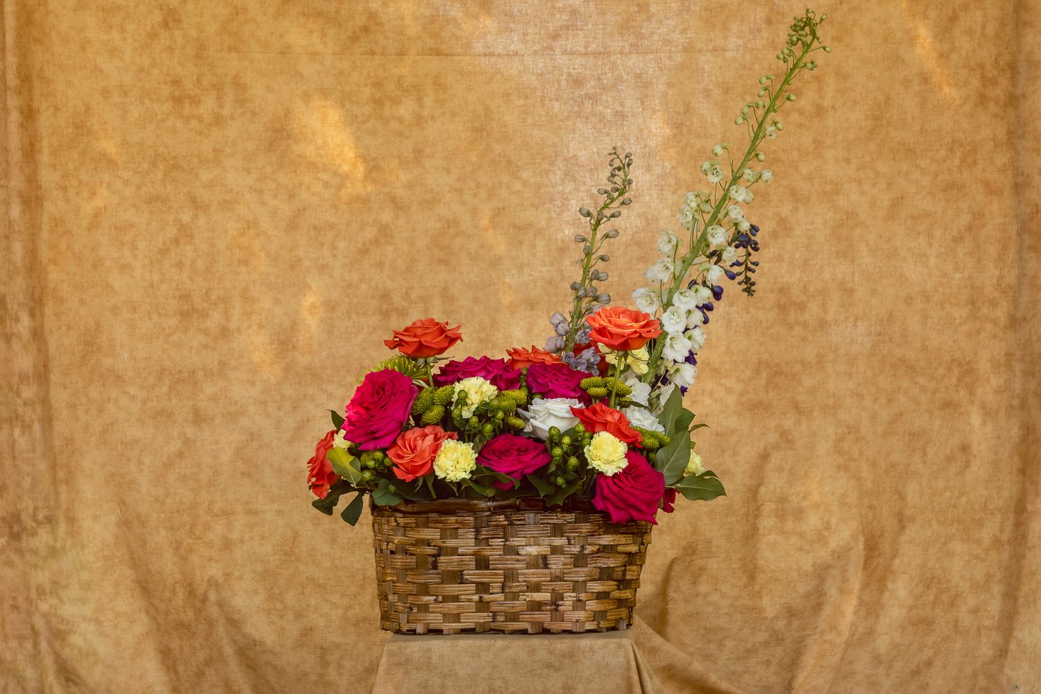 Vibrant floral arrangement in a wicker basket featuring red, orange, and yellow roses with blue accent flowers on a tan background.