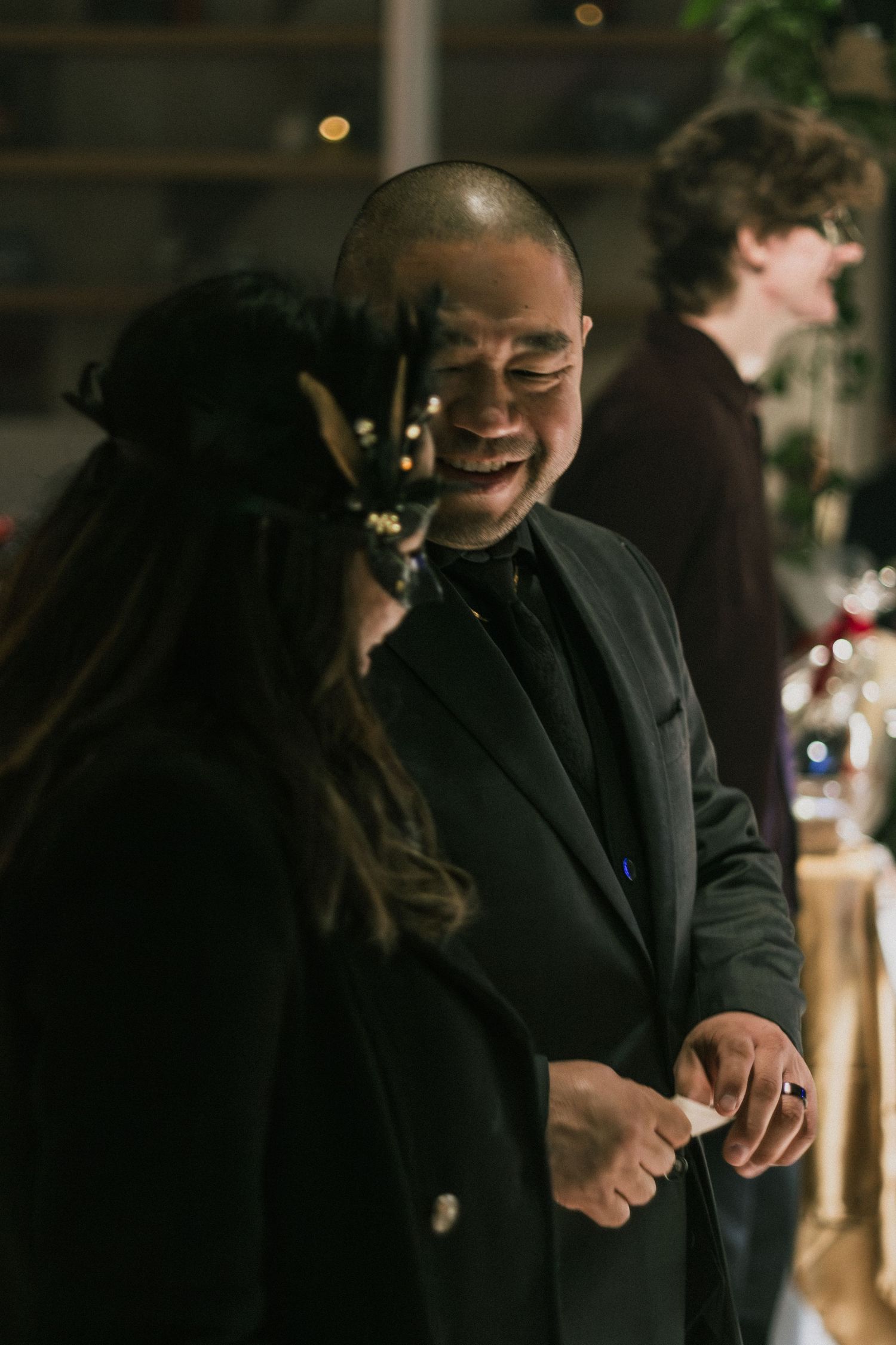 A smiling man in a black suit laughs with a woman wearing a decorative headpiece at an indoor evening event.